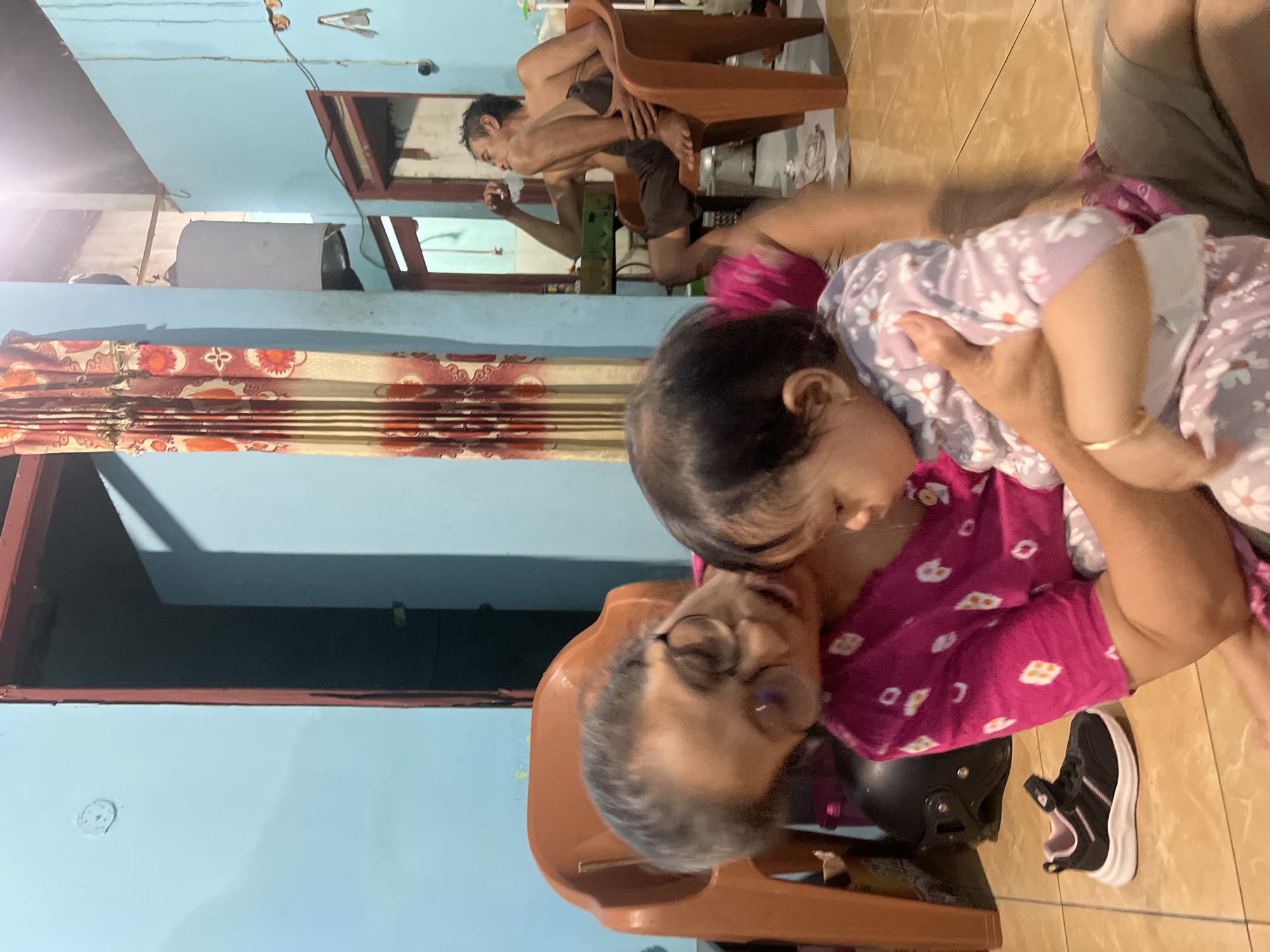 In a domestic indoor setting, likely in Kecamatan Pati, Indonesia, a woman with graying hair and glasses is holding a baby. The woman is wearing a pink patterned shirt, and the baby is dressed in a light-colored floral outfit. In the background, another person, a man, is seated in an orange chair, appearing to be smoking or drinking. The room is painted light blue, with a red-trimmed doorway visible. A patterned curtain hangs on the right side of the frame. On the floor, a black helmet and a child's sneaker are visible. The lighting suggests an indoor environment, possibly in the evening or with artificial light sources.