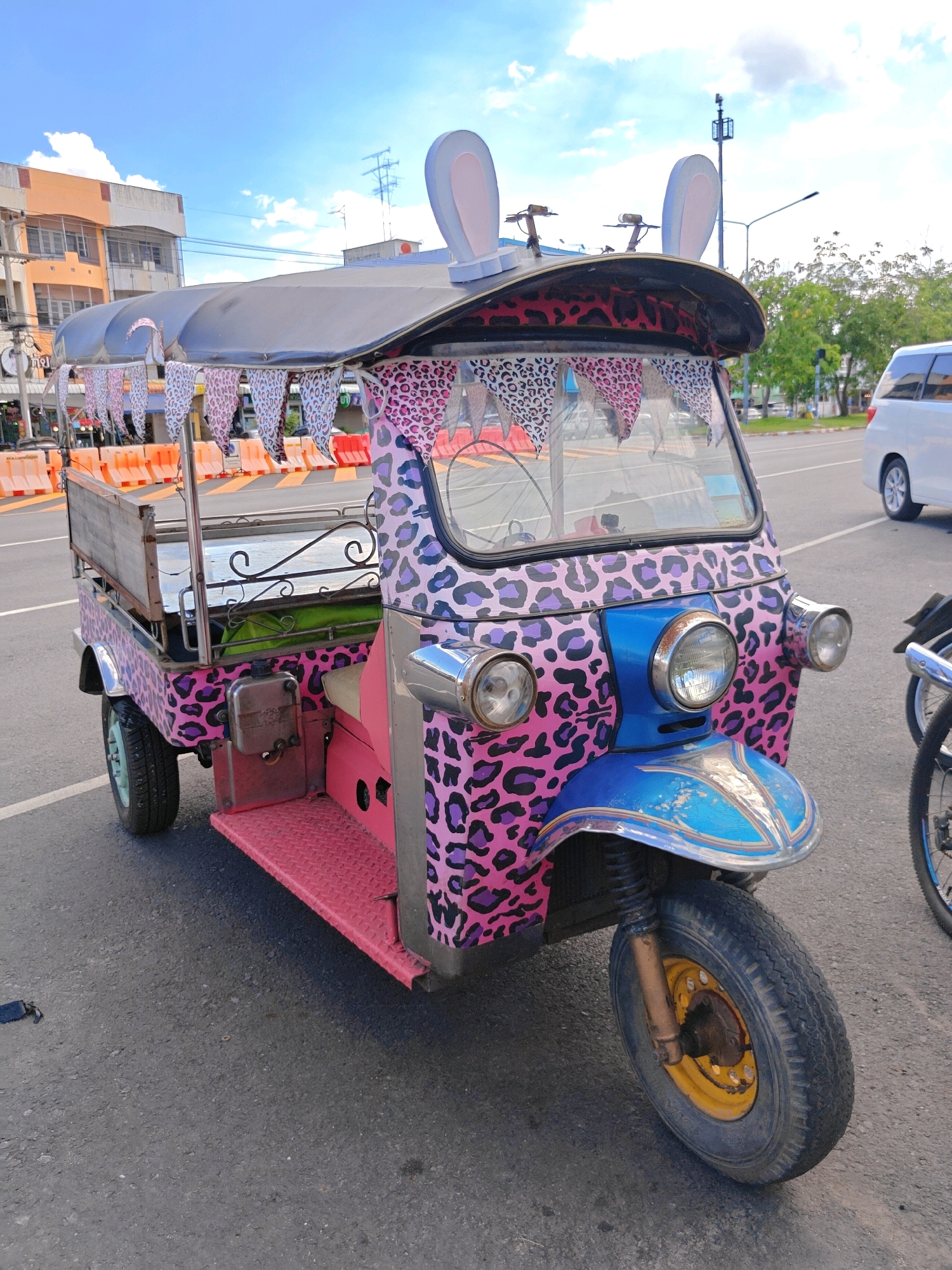 A pink and purple leopard print decorated three-wheeled auto rickshaw, commonly known as a tuk-tuk, is parked on a paved road. The tuk-tuk features large, white bunny ears on its roof and a row of decorative triangular flags strung along the front. The vehicle is brightly colored, with pink, blue, and silver accents. The front of the tuk-tuk has two round headlights. The background shows a street scene with buildings, trees, and a white van. Orange traffic barriers are visible on the left side of the image. The sky is blue with scattered clouds. The image appears to have been taken in Phra Nakhon Si Ayutthaya City Municipality, Thailand, based on the architectural style of the buildings and the presence of this distinctive mode of transportation.