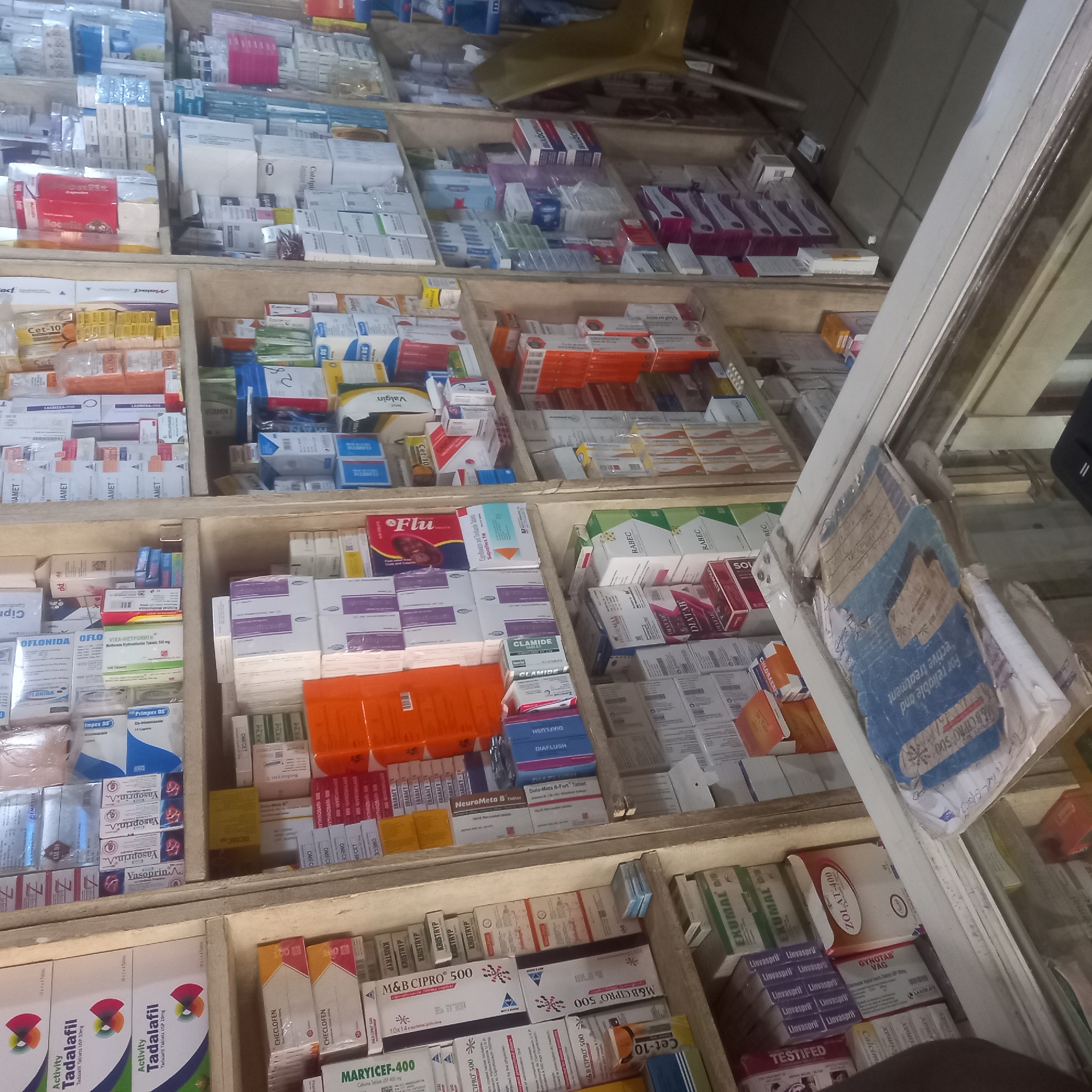 This image offers an eye-level, cluttered view inside a pharmacy or drug store in Maiburiji, Nigeria. The scene is dominated by numerous pharmaceutical products, primarily medications in various boxes and packets, stacked on wooden shelves. No people are visible, focusing attention entirely on the inventory.
The shelves are densely packed, with products arranged both vertically and horizontally, creating a busy yet functional display. The lighting suggests an indoor setting, likely with artificial light. A worn, blue folder or document hangs partially visible on the right side of the frame, near a white shelving upright.
Numerous brand and generic drug names are legible, including "Activity Tadalafil Tablets USP 20mg," "CHECLOFEN," "MARYICEF-400," "Vasoprin," "OFLONIDA," "VIXA-METFORMIN 500 mg," "OMECE," "ERYTHROBAR-500," "KRISTRYP," and "M&B CIPRO 500," which is explicitly described as "For reliable and effective treatment" of "Ciprofloxacin 500mg." Other identifiable texts include "Cet-10," "FRABACT," "IAMET," "NeuroMeta B," "Dolo-Mets B-Fort Tablet," "DIAFLUSH," "EXUMAL," "ZOLAT-400," "Linvaspril," "GYNOTAB VAG," "TESTIFED," "CLAMIDE," "Flu," "Suprafix TH," "Maloct," "DIATAB," "Valgin," "RABEC," "SOLMATE," and "Glaformir." The image conveys the impression of a well-stocked, local pharmaceutical outlet.