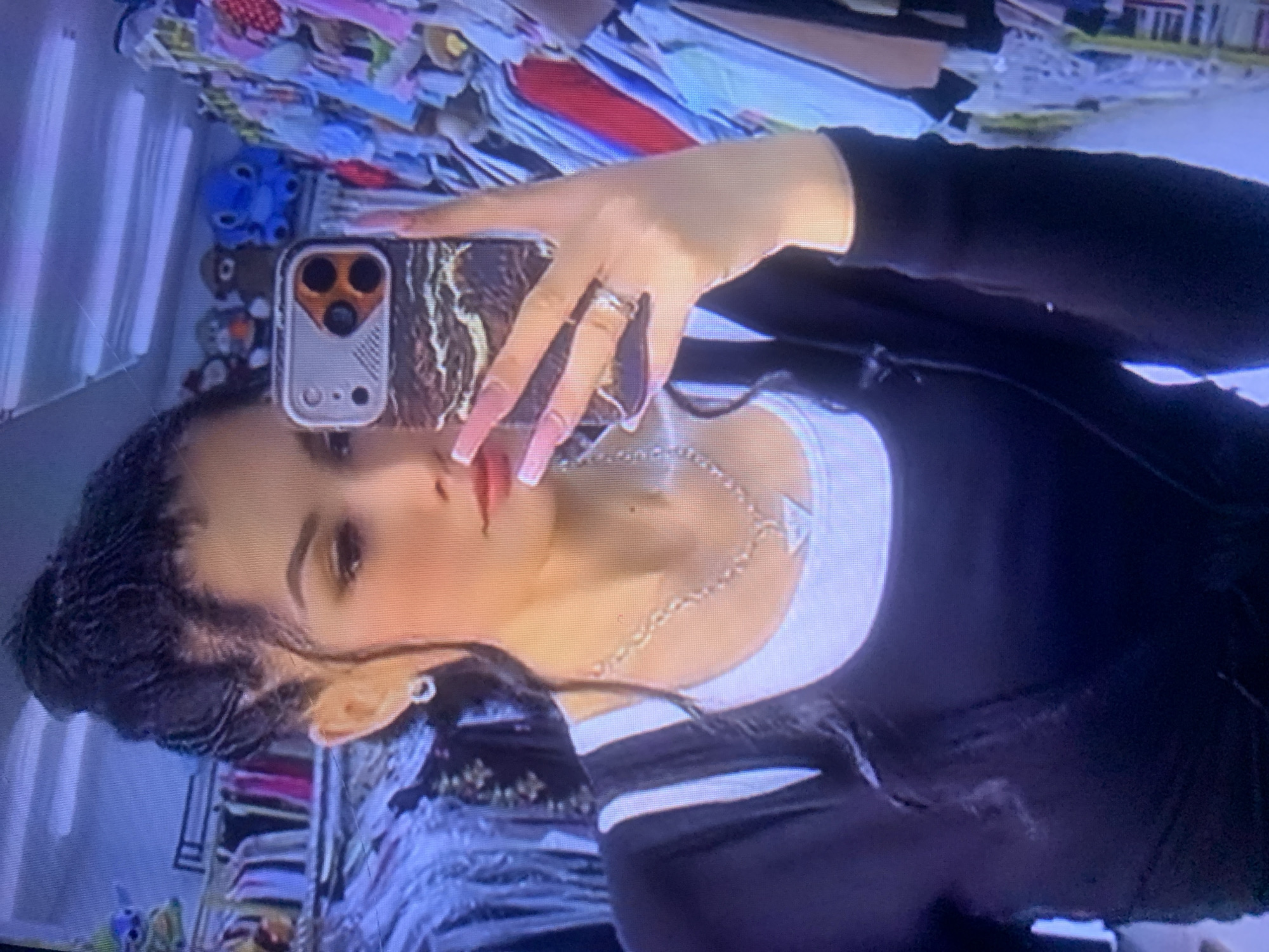 The image shows a young woman with dark hair pulled up in a bun, taking a selfie. She is wearing a black long-sleeved shirt over a white tank top, a silver necklace with a triangular pendant, and a ring on her left hand. She has long, light pink acrylic nails. In the background, clothing racks filled with colorful garments are visible, suggesting she is in a retail store. The scene does not provide any specific visual cues to identify the location as Almaty, Kazakhstan.