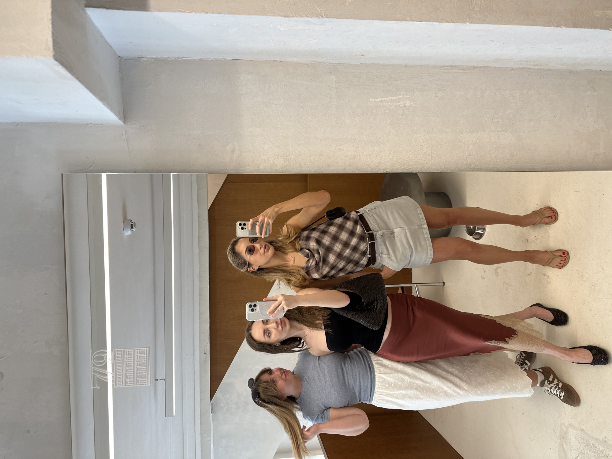 Three young women are taking a mirror selfie in what appears to be a retail space or a cafe in Moscow, Russia. The woman on the left has her hair pulled up and is wearing a grey t-shirt and a long white lace skirt. The woman in the middle is wearing a black tube top, a reddish-brown satin skirt, and holding a black purse. The woman on the right is wearing a plaid camisole, a light denim skirt, and sunglasses. They are all looking at their phones, which are held up to take the photo. The background shows a white wall with some wooden accents, a large mirror reflecting them, and a logo that appears to say "70" with a building design. The floor is a light color, and there are some light fixtures on the ceiling.