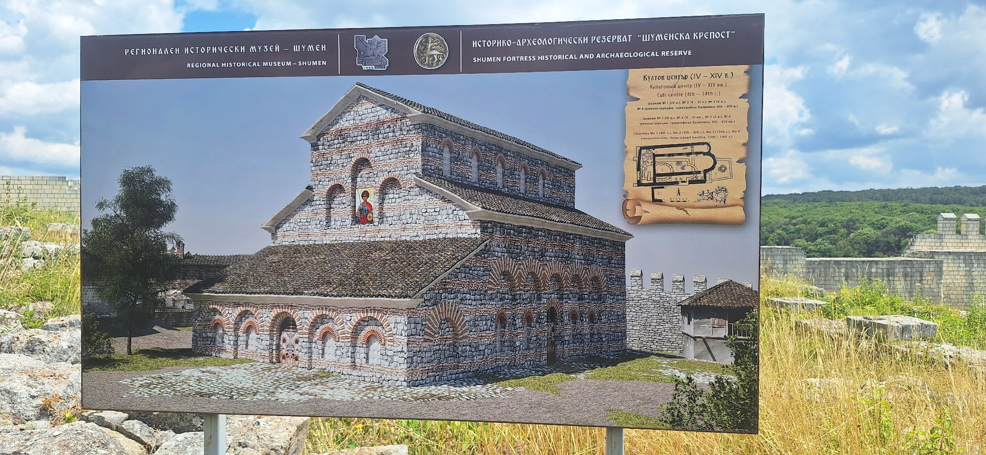 The image displays an informational sign in front of an outdoor historical site. The sign features a detailed illustration of an ancient stone building, possibly a church or basilica, with arched doorways and windows, and a tiled roof. Above the illustration, text in both Cyrillic and Latin scripts identifies the location as the "Regional Historical Museum – Shumen" and the "Shumen Fortress Historical and Archaeological Reserve." To the right of the illustration, a schematic drawing of the building's floor plan is shown on a scroll-like background, with accompanying text in Cyrillic and Latin detailing historical periods and structures within the reserve, including "Cult centre (4th – 14th c.)" and "Churches No. 1 (4th c.), No. 2 (5th – 6th c.), No. 3 (10th c.), No. 4 (reconstruction, Three-naved basilica, 13th – 14th c.)."
The background behind the sign shows the actual ruins of the historical site, with stone walls and scattered blocks overgrown with grass and wildflowers. A dense green forest stretches across the horizon. The sky is partly cloudy. The sign is supported by a metal pole, and the foreground consists of rough terrain with large rocks and tall, dry grass.