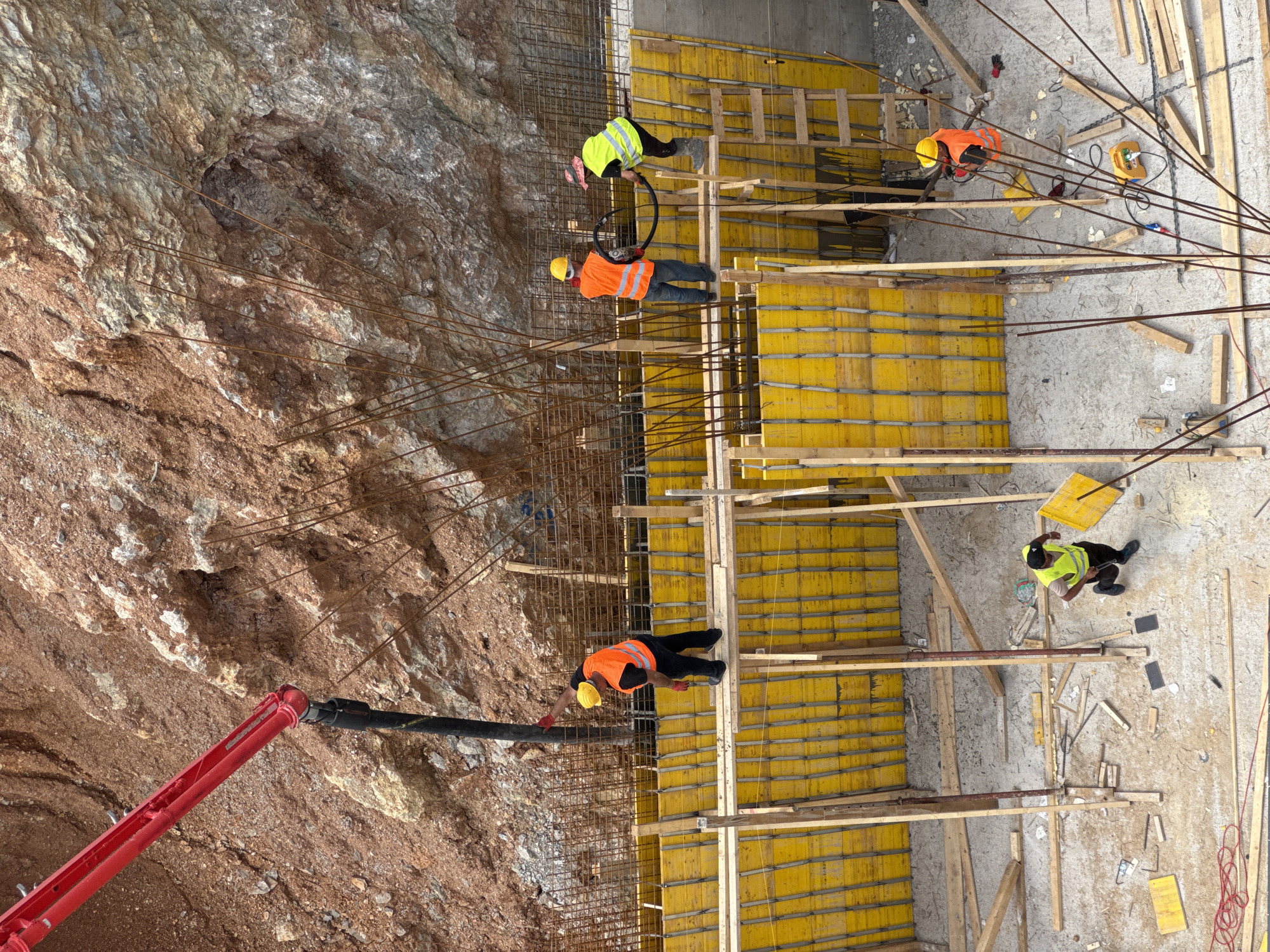 A construction site is depicted in an overhead shot, potentially in or near Budva, Montenegro. A rock face forms one side of the construction, with a red concrete pump arm extending from the lower left towards the rock. Workers wearing hard hats and safety vests are positioned amidst a steel rebar structure and yellow formwork. Various tools and materials are scattered around the site.