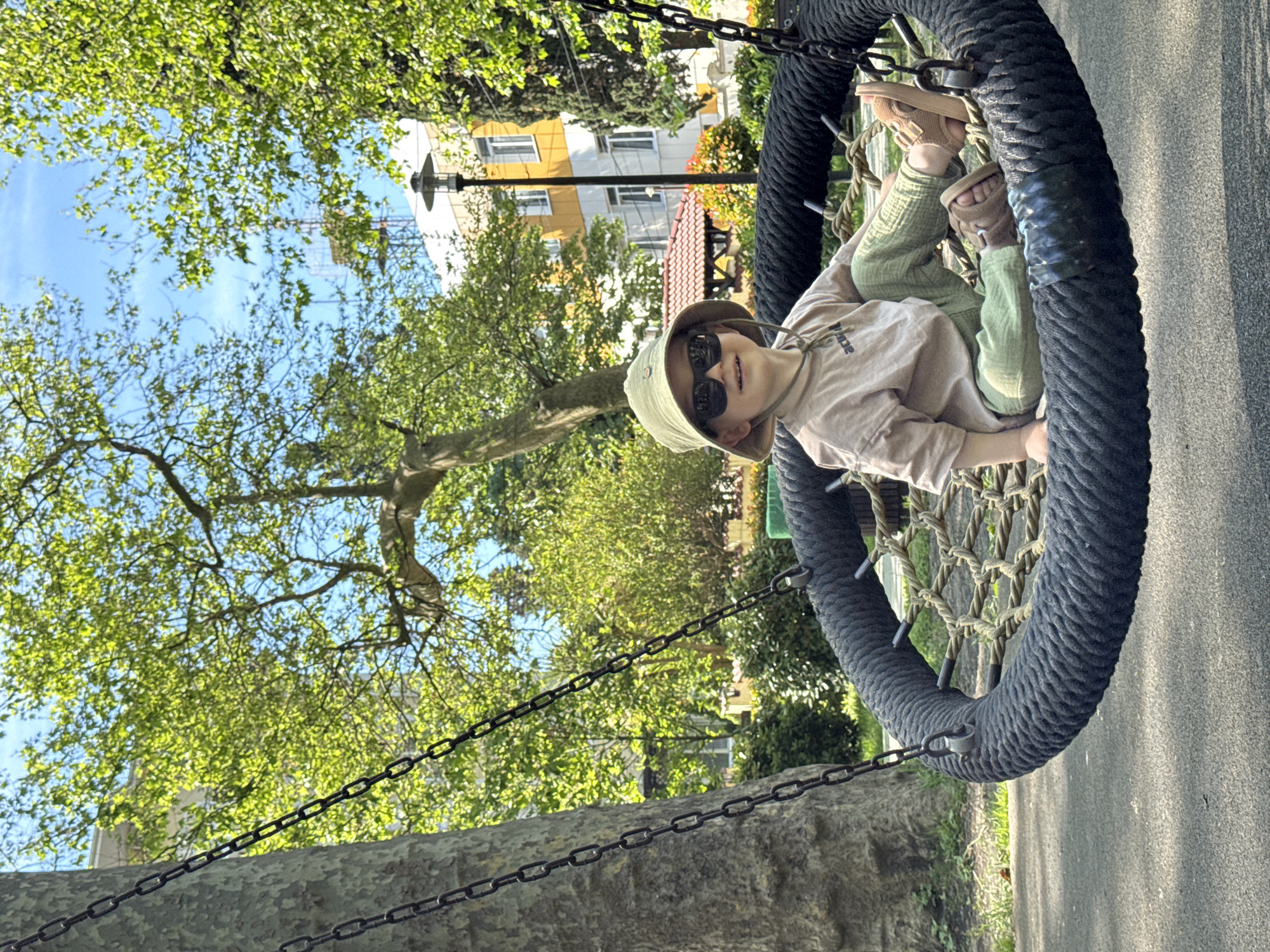A young child wearing a khaki bucket hat and sunglasses sits in a circular swing at what appears to be a park or playground. The swing is made of rope and has a thick, black, rope-covered rim. Chains attach the swing to a tree. The background includes green trees and a multi-story building with yellow and white sections. The location may be in Sochi, Russia.