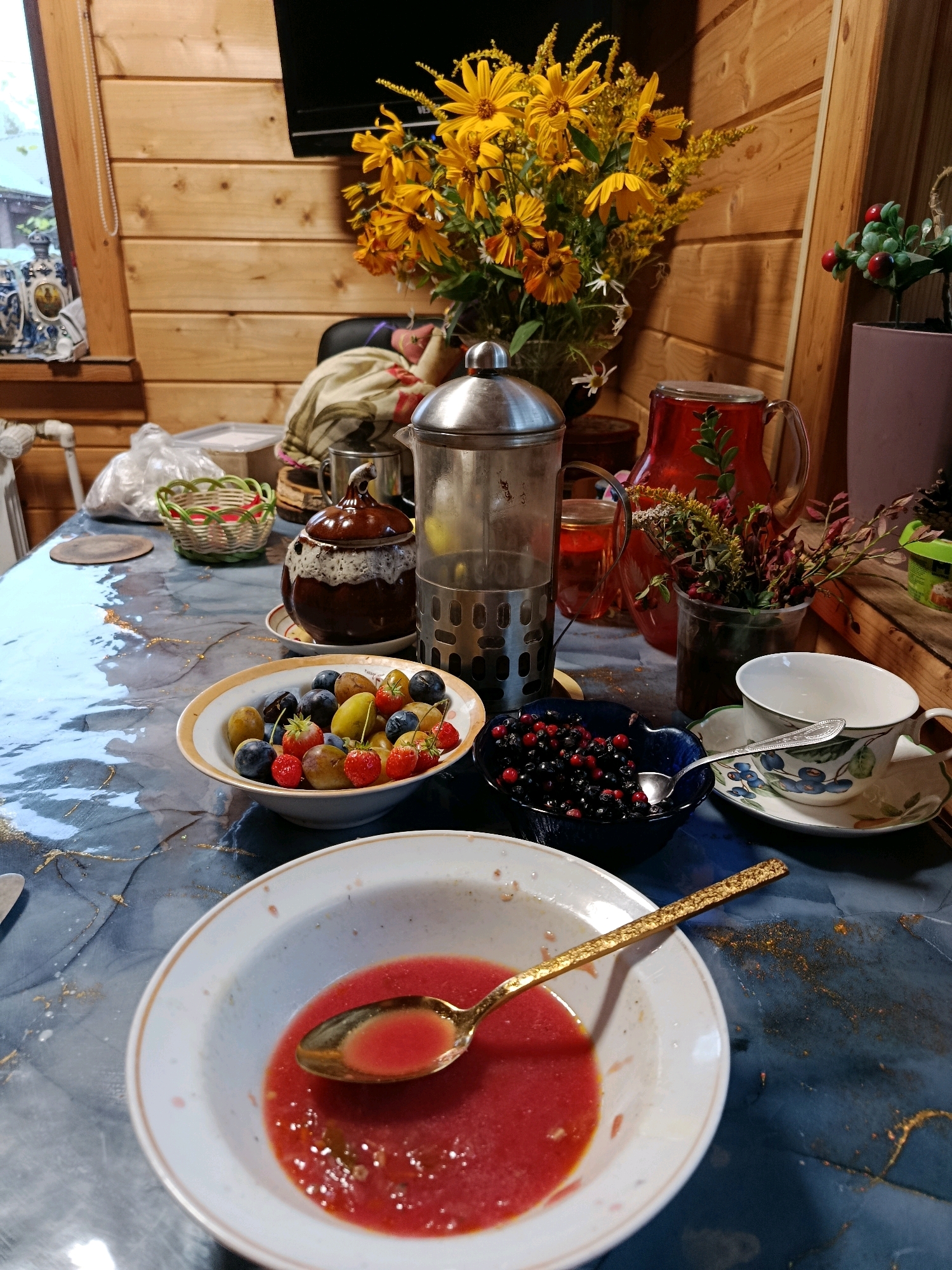The image depicts a table laden with food and drinks, possibly a breakfast or snack spread, set against a backdrop of wooden paneling. The scene is arranged on a dark blue tablecloth with a marbled pattern and gold accents.
In the foreground, a white ceramic bowl contains a portion of what appears to be a red liquid or purée, possibly tomato soup or gazpacho, with a golden spoon resting inside. To the left of this bowl is another white bowl with a gold rim, filled with a mixture of berries, including blueberries, strawberries, and possibly grapes or plums.
To the right, a dark blue bowl holds a generous serving of blueberries and redcurrants, with a silver spoon visible inside. Beside it sits a delicate teacup and saucer with a floral pattern featuring blue flowers and green leaves.
Behind these bowls, various items contribute to the rustic and inviting atmosphere. A glass French press coffee maker stands prominently. To its left, a brown ceramic pot with a patterned lid is visible. A red glass pitcher, filled with a red beverage and garnished with a sprig of greenery, is positioned behind the French press. A small arrangement of dried flowers and twigs is in a clear plastic cup next to the pitcher.
In the background, a bouquet of bright yellow flowers with hints of orange and white, possibly sunflowers or similar blooms, is displayed in a vase. Further back, a woven basket and a plastic-wrapped item are partially visible, suggesting other food items or supplies. The wooden walls and a window with a glimpse of greenery complete the setting, evoking a sense of a cozy, perhaps rural or cottage-like environment.