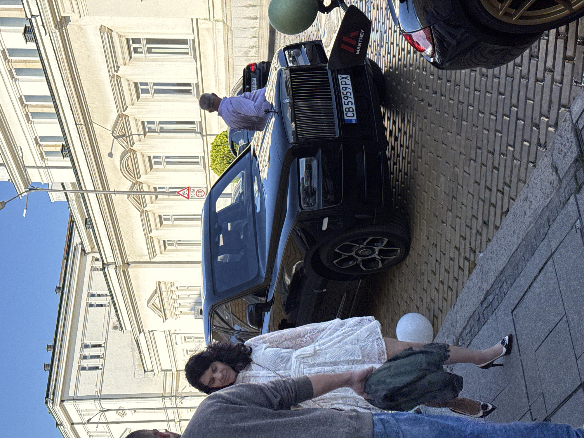 The image shows a street scene in Sofia, Bulgaria, with a Rolls Royce parked on a cobbled street near a building with a light facade. A man in a light purple shirt is leaning on the hood of the car, possibly inspecting it. A woman in a white lace dress and a man in a grey shirt and blue jeans are on the sidewalk near the car. The woman is lying down, seemingly unconscious or in distress, with her legs extending towards the sidewalk edge. She is wearing high heels. The man in grey is kneeling next to her, possibly providing assistance. The car has a license plate "CB 5959 PX" and a "Manthey" sign on the hood. A white sphere is on the sidewalk. A street sign "30 HA" is also visible near the building.