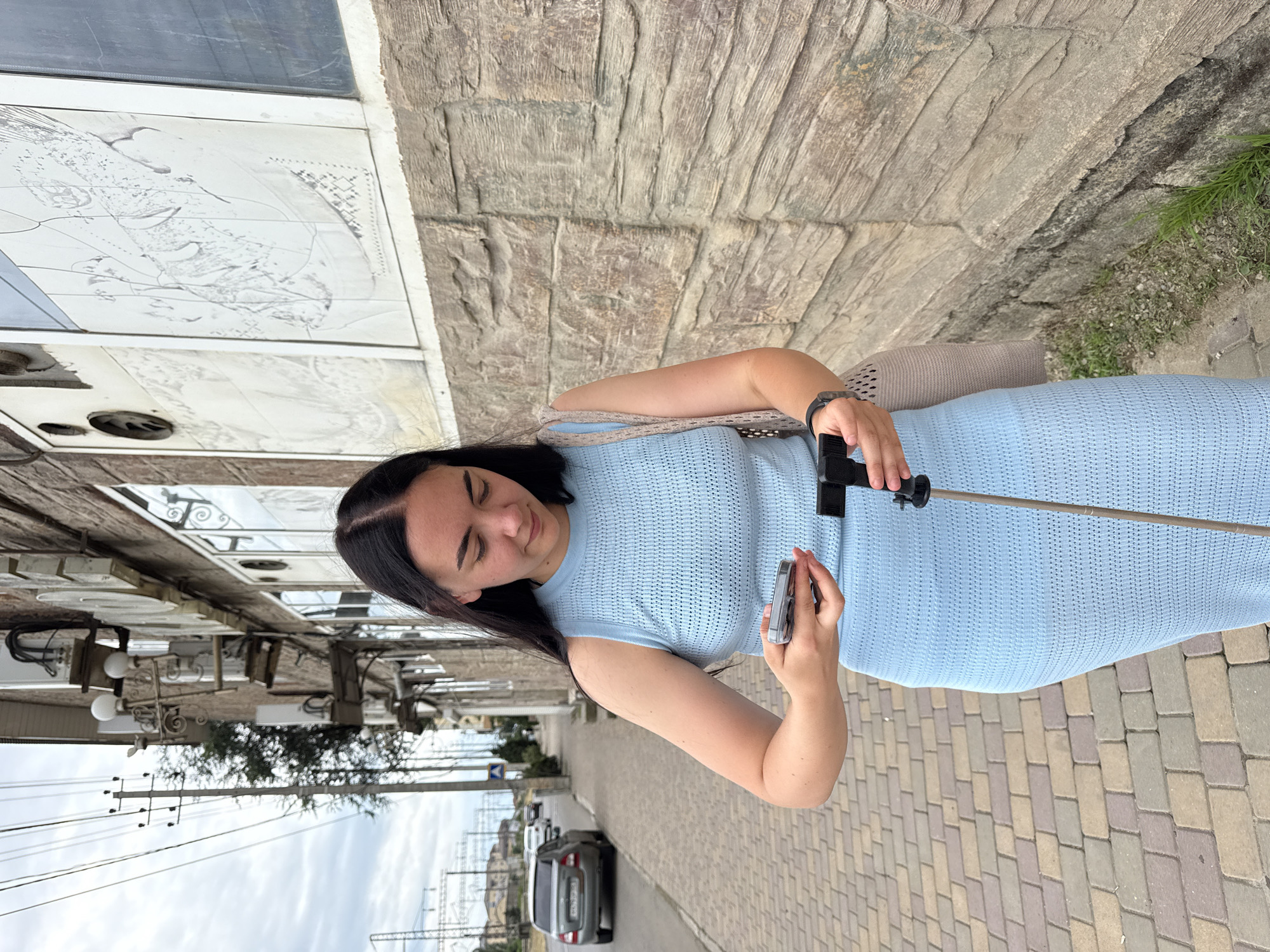 A young woman with dark hair is taking a selfie with a smartphone attached to a selfie stick. She is wearing a light blue knitted dress and a beige bag is slung over her shoulder. She is standing on a cobblestone sidewalk next to a building with a stone facade and large windows that reflect the surroundings. The woman has a slight smile on her face. In the background, there is a street with parked cars and utility poles with wires. The scene appears to be outdoors in daylight, with an overcast sky. The presence of cobblestone streets and the architectural style of the building suggest an urban or historic setting. The text "Derbent, Russia" is provided as location context.