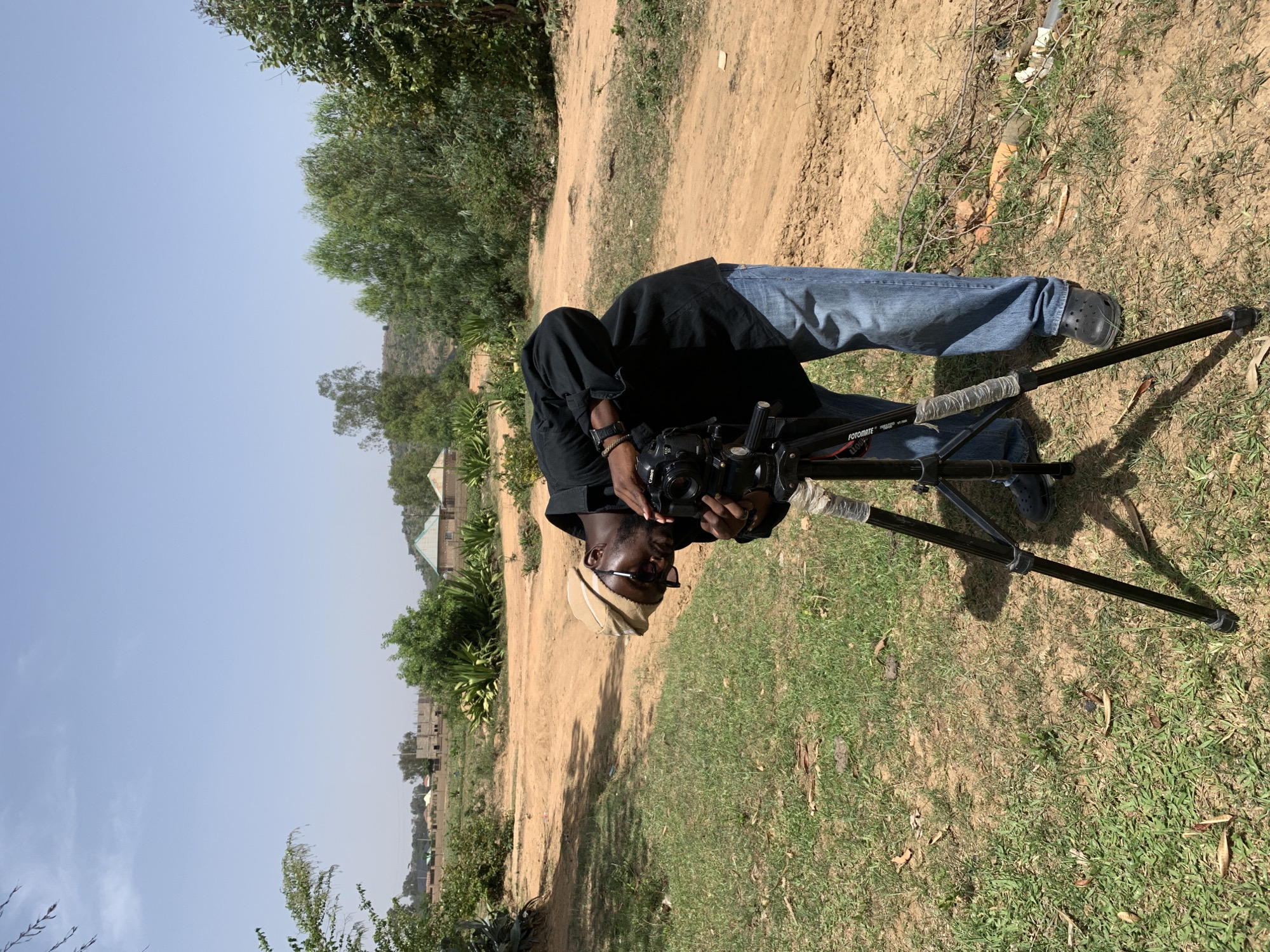 A man stands outdoors in Gwafan, Nigeria, operating a camera on a tripod. He wears a black shirt, blue jeans, dark shoes, a light-colored hat, and glasses. The ground is a mix of grass and dirt. In the background are buildings and trees under a light blue sky.