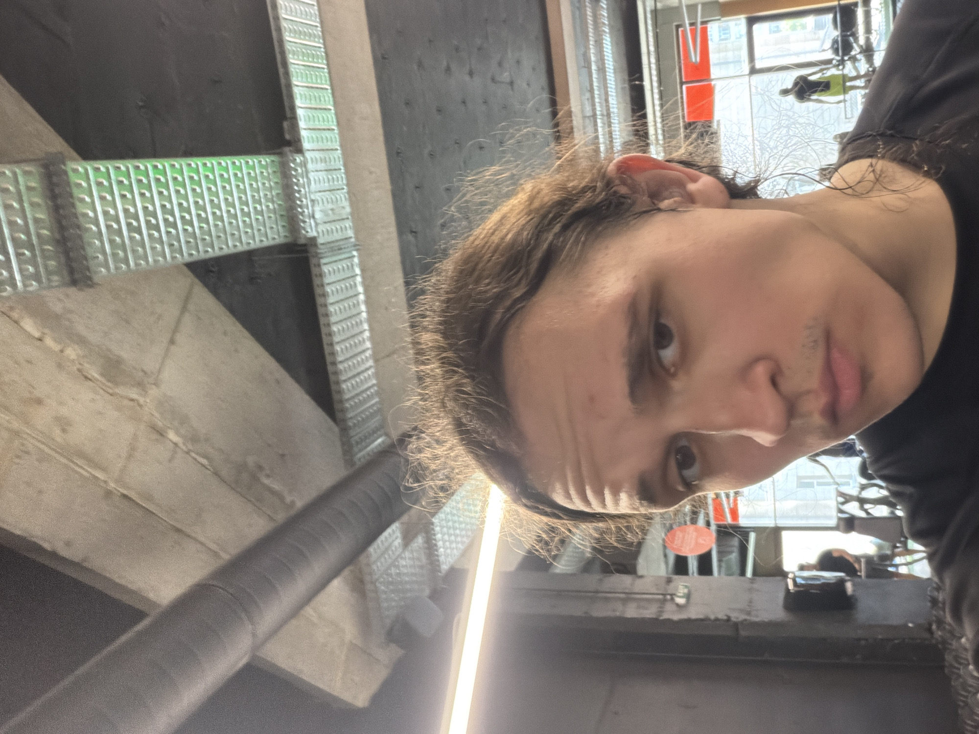 The image shows a selfie taken from a low angle. A person with long, brown hair is looking into the camera. The background is an industrial or commercial setting, possibly a gym or a similar recreational facility. The background includes metal beams, ductwork, and window views of an urban landscape. Through the windows, there are blurred views of buildings and possibly other people. The location of this photo cannot be determined as the background does not have any recognizable landmarks of Plovdiv, Bulgaria.