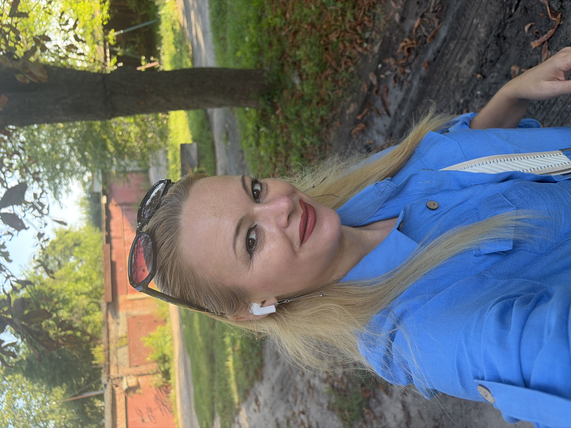A blonde woman with sunglasses perched on her head and white earbuds in her ears is captured in a selfie. She is wearing a blue collared shirt with the sleeves rolled up and fastened with buttons. Her hair is light blonde and falls past her shoulders. She is wearing lipstick and has light makeup. The background consists of a dirt path, grass, and trees, suggesting an outdoor setting. A brick wall and garage doors are visible in the distance.
