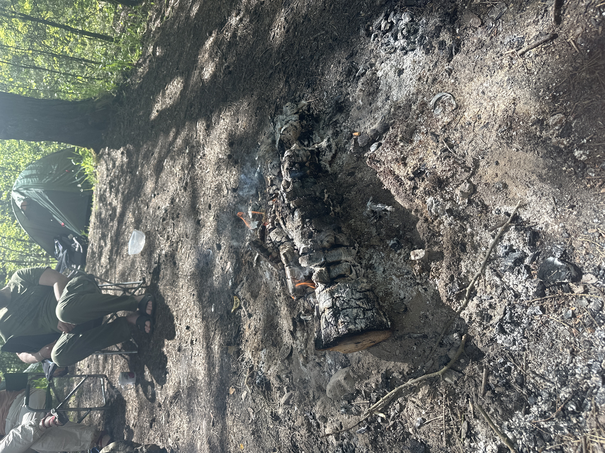 A ground-level, rotated view shows the aftermath of a campfire in what appears to be a wooded area in or near the Юзуфоўскі сельскі Савет in Belarus. The fire pit contains burnt logs, embers, ash, and charred debris. Some residual smoke is visible. To the left, two people are seated in folding chairs. A tent is partially visible in the background, suggesting a campsite. The ground is covered in dirt, leaves, and small branches.