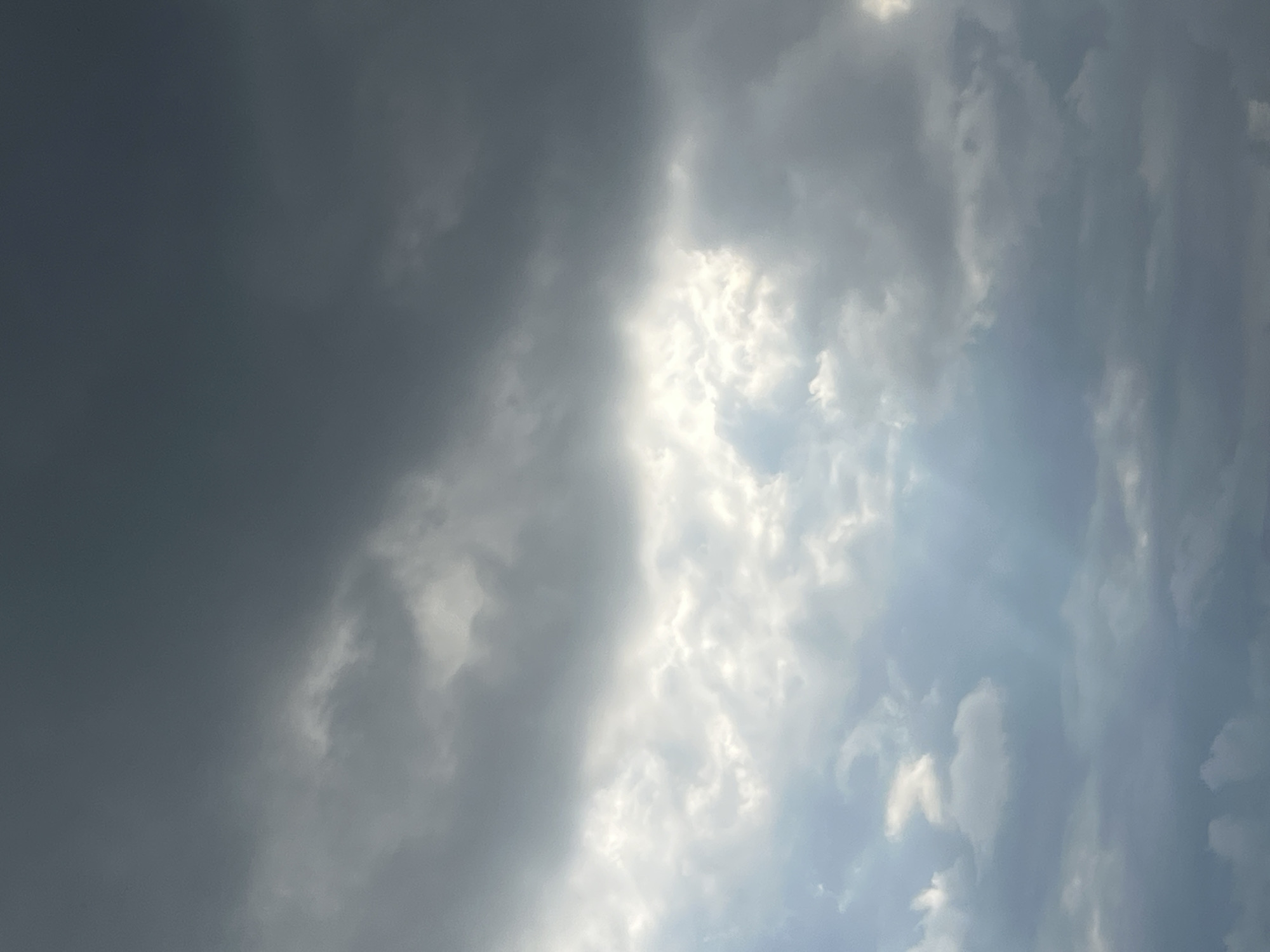 The image shows a stormy sky with dark, heavy clouds dominating the upper portion. In the middle of the frame, a break in the clouds allows sunlight to stream through, illuminating some of the lower clouds with a bright, ethereal glow. The light creates a dramatic contrast with the darker, more ominous clouds above and around it. The lower part of the image is also filled with clouds, some appearing lighter and more diffused, while others are darker and more defined. There are no people, buildings, or any other objects visible in the frame; the image solely captures the atmospheric conditions of the sky. The specific location in Chelyabinsk, Russia, cannot be determined from the visual information presented.