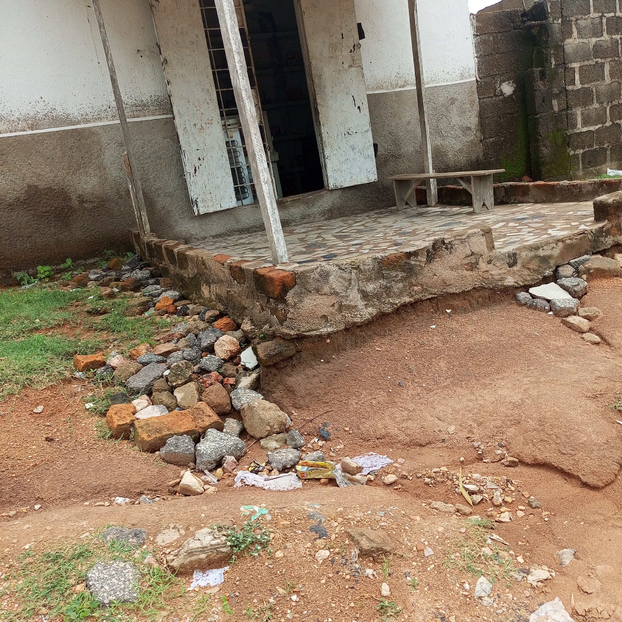 This image depicts an outdoor scene in Jalingo, Nigeria, likely in a somewhat dilapidated area. The primary subject is a weathered building with large, white, shuttered windows and a veranda supported by simple poles. The building's facade is a mix of white and textured gray, showing signs of age and wear. In front of the building, a paved patio area with a patterned tile design is visible, leading to a rough, unfinished concrete foundation. To the right, a section of a brick wall, weathered and partially covered in moss, stands prominently.
In the foreground, the ground transitions from grass and scattered debris to loose, reddish-brown soil. A collection of rocks and broken bricks are piled near the foundation of the building, suggesting some form of construction or decay. Litter, including plastic and paper, is scattered across the ground, particularly in the reddish soil area. A simple, wooden bench with angled supports sits on the paved patio, facing away from the viewer and towards the brick wall.
The scene appears to be set during daylight, though the sky is not visible, the lighting suggests an overcast or shaded day. The overall impression is one of disrepair and neglect, with elements of nature beginning to reclaim the man-made structures. There is no discernible activity or event taking place, and no text is visible in the image. The location cues are general, pointing to a rural or semi-urban environment in Nigeria.