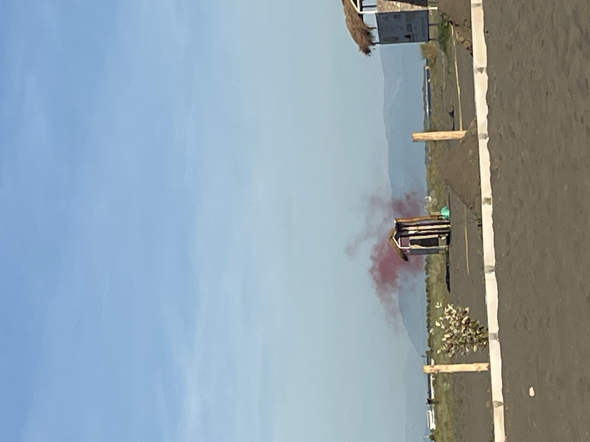 The image depicts an outdoor scene with a clear blue sky. In the mid-ground, there is a small, dark wooden structure with a pitched roof, resembling a shed or a toilet. Red smoke is emanating from behind and above this structure, suggesting it might be a signal or a deliberate emission. To the right of the shed, a larger structure with a thatched roof is partially visible, appearing to be a covered seating area or a kiosk. In the foreground, a dark sandy or dirt surface stretches across the frame, with a white concrete curb running along the bottom edge. A thin, leafless shrub grows near a wooden post in the mid-ground. In the background, distant mountains or hills are visible under a hazy sky. The overall environment appears to be a coastal or open area, possibly a beach or a park. There are no identifiable people or animals in the scene. The location is identified as the city of Pistula, Montenegro.