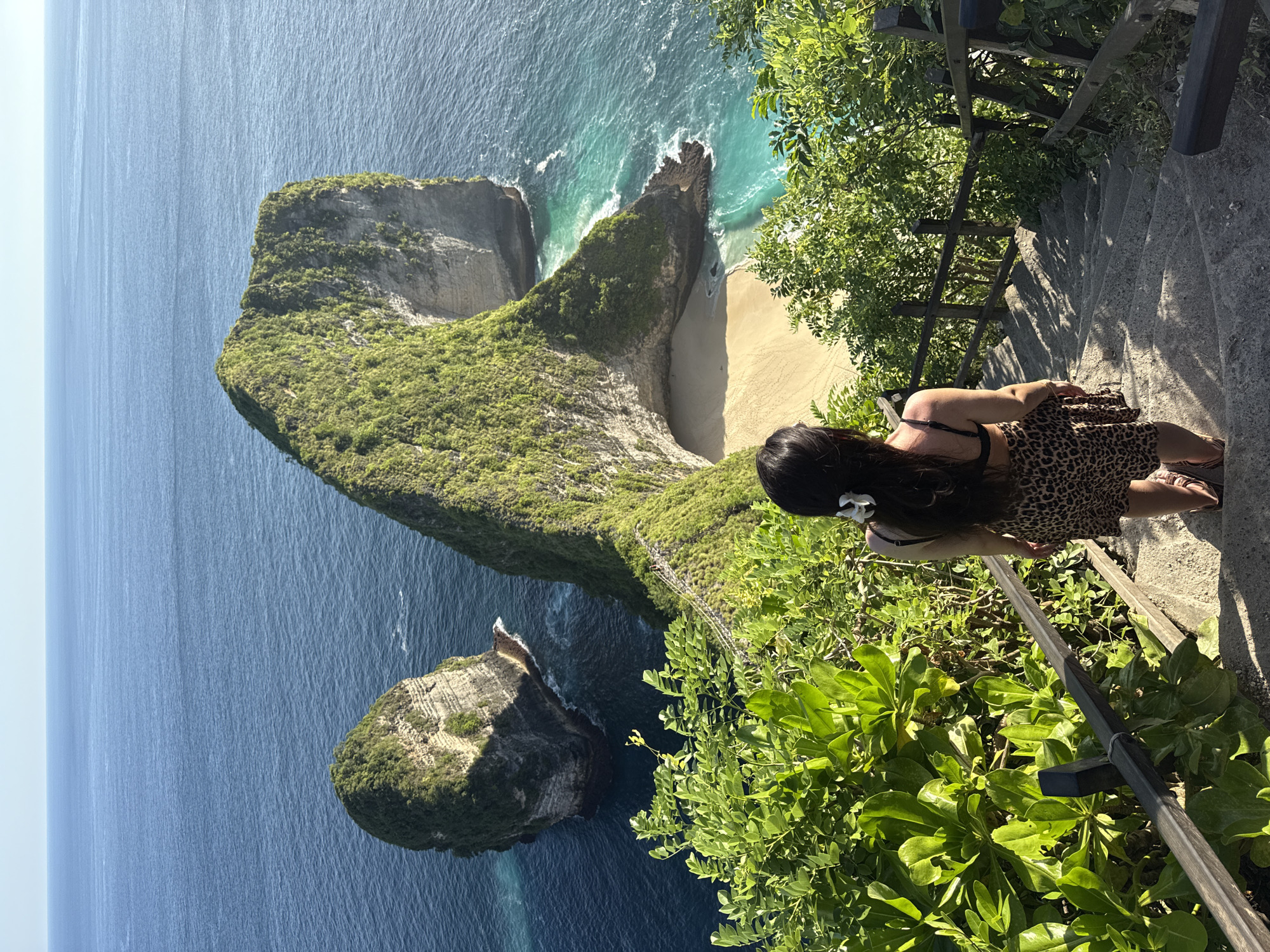 A vertical full shot depicts a woman standing on a staircase, looking down at a scenic view of the coastline and ocean in Nusa Penida, Indonesia. The woman has long dark hair with a white flower tucked behind her ear, and she is wearing a leopard print dress and sandals. She stands on concrete steps, next to a wooden railing and lush green plants. To her left, the coastline features a large, green, T-shaped rock formation that extends into the turquoise ocean, revealing a sandy beach. Beyond this formation, the deep blue ocean stretches out to the horizon under a clear sky.