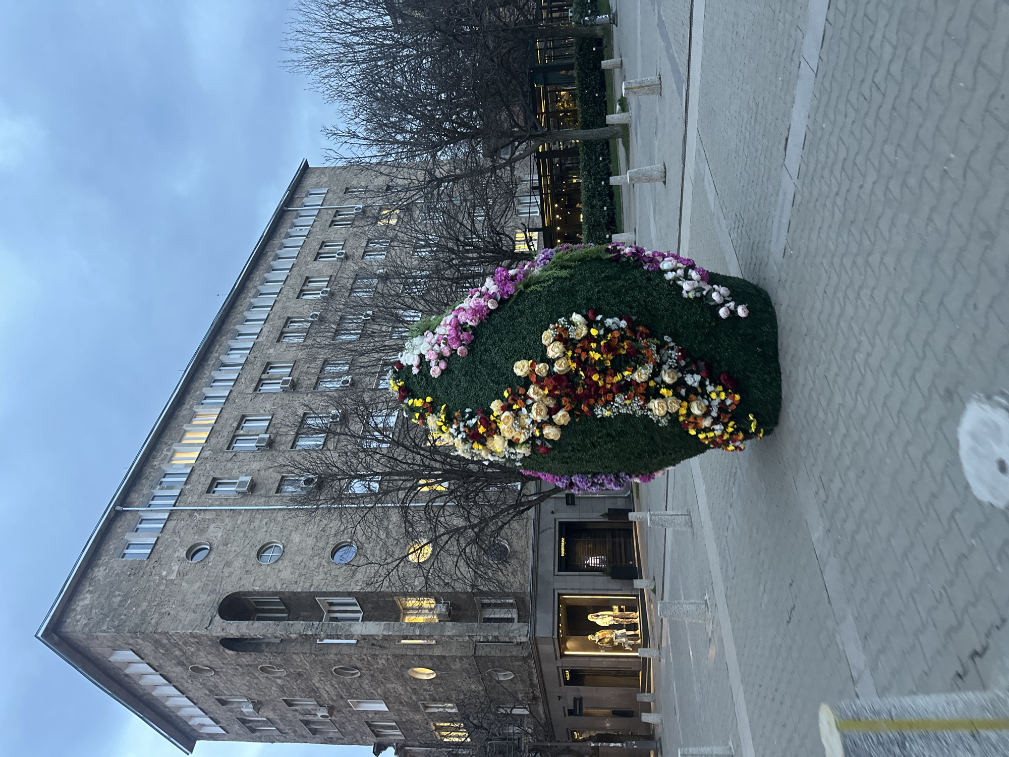The image depicts a large, egg-shaped floral arrangement positioned on a paved area in an urban setting. The arrangement is predominantly green, adorned with flowers in shades of pink, white, yellow, and red. Behind the floral arrangement is a large, grey, multi-story building with rectangular and circular windows. Bare trees are visible in front of the building. A store front can be seen at the base of the building. The sky is overcast. The paving consists of grey blockwork. The location is the city of Sofia, Bulgaria.