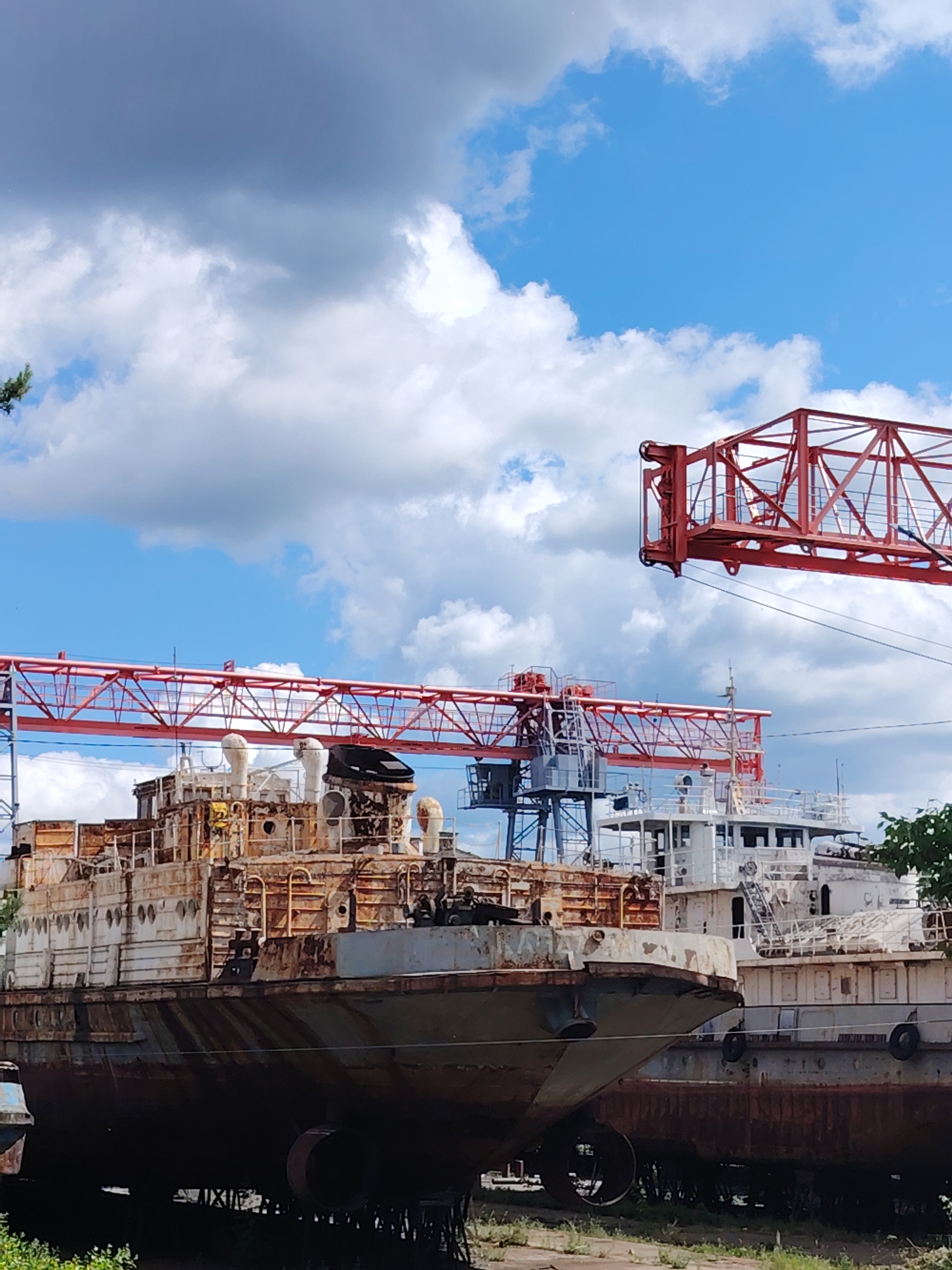 The image depicts a shipyard in Chaikovsky, Russia, featuring several decommissioned ships resting on land. The primary vessel in the foreground is heavily rusted and appears to be in a state of disrepair. Behind it, partially visible, are other ships in similar condition, some painted white. A large red crane structure looms over the scene. The sky is blue with scattered white clouds. The ships rest on some sort of supports or scaffolding in an area overgrown with vegetation.