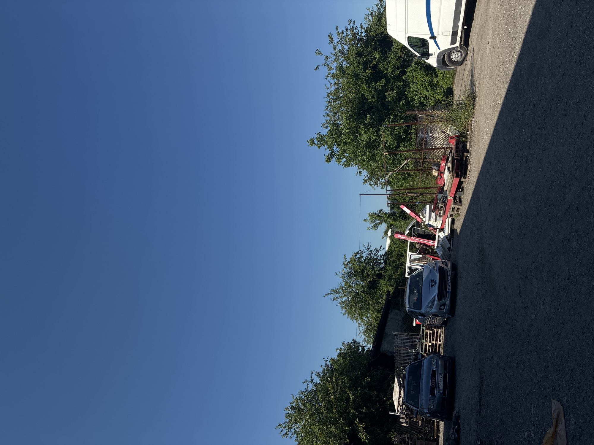 A grey Audi A4 sedan with license plate BT 0237 KH and a silver Peugeot 208 with license plate P 8997 KX are parked in a gravel lot under a clear blue sky. To the left of the vehicles, stacks of wooden pallets and some metal fencing are visible. Behind the cars, a low-slung building with a dark roof is partially obscured by trees. In the background, more trees and foliage create a green backdrop. To the right, a white van with a blue stripe is parked. Various metal structures, red and white equipment, and more pallets are scattered around the area. The scene appears to be an outdoor storage or industrial yard.