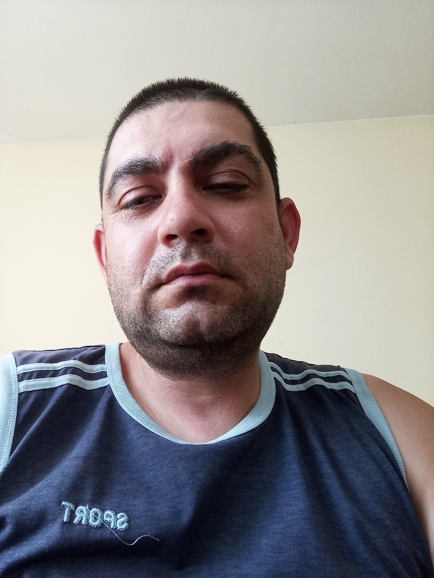 A close-up, eye-level portrait features a man indoors in Ravnets, Bulgaria. He appears to be middle-aged, with short, dark, closely-cropped hair and dark stubble or a short beard covering his jawline. His face is full, and he looks directly at the camera with a neutral, somewhat serious or slightly weary expression, his eyes appearing heavy-lidded.
The man is wearing a dark blue sleeveless shirt, or tank top, with light blue trim around the neck and armholes. Three distinct light blue horizontal stripes are visible on each shoulder of the garment. On the lower front of the shirt, the word "SPORT" is embroidered in light blue thread, appearing mirrored in the image, suggesting the photo was taken with a front-facing camera. A single loose thread hangs from the embroidery near the text.
The background consists of a plain, light beige or off-white wall, indicating an indoor setting. The lighting is generally even, illuminating his face. No other specific location cues or objects are visible in the frame, maintaining focus on the subject.