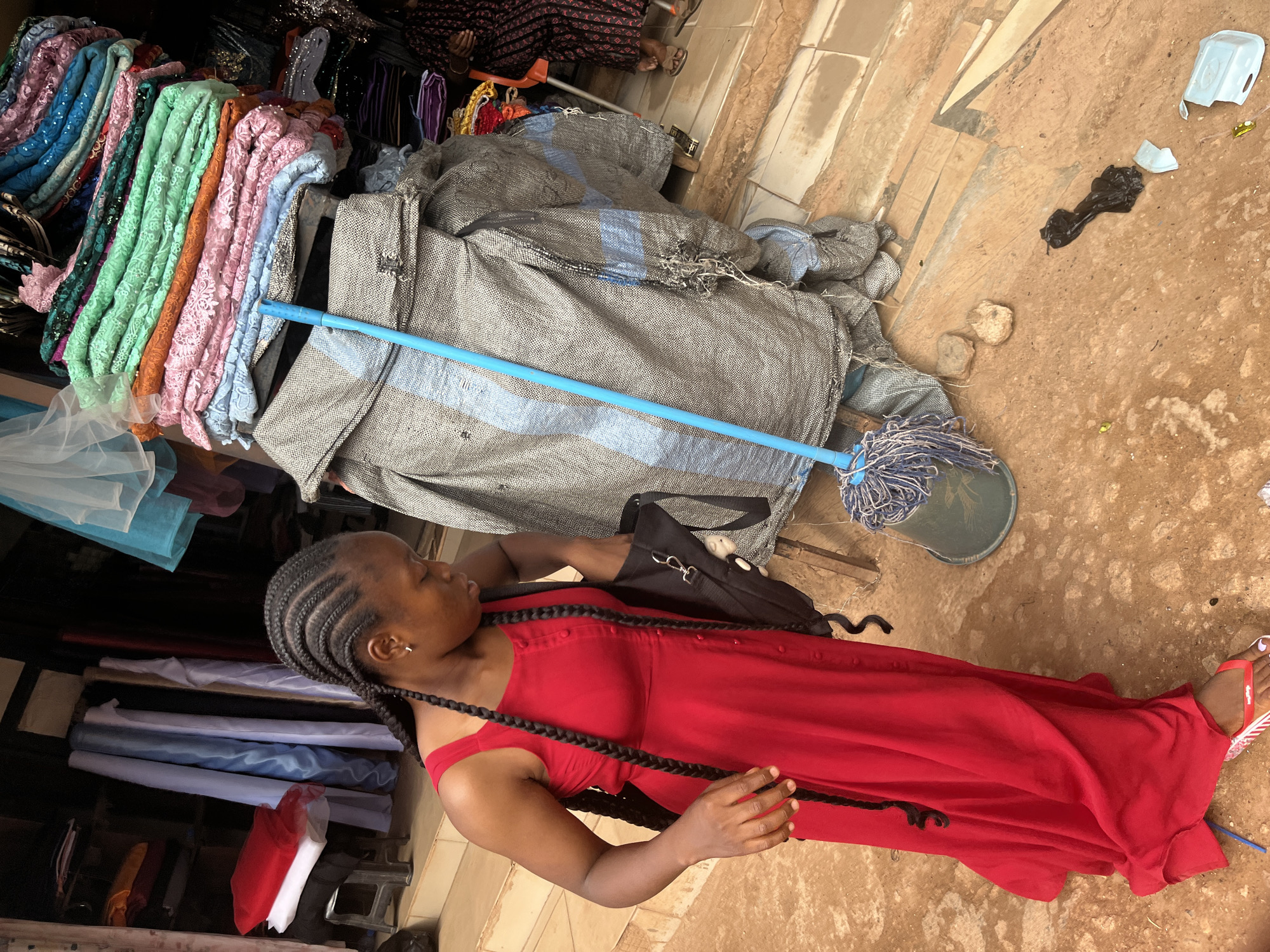In Jos, Nigeria, a woman with braided hair, wearing a sleeveless red dress, is seen in a marketplace. She is reaching into a black bag that hangs from her shoulder. In the background, stacks of colorful fabrics are displayed, and a mop with a blue handle rests against a large, tarp-covered object. The ground is a mix of dirt and tiled sections, with some debris scattered around.