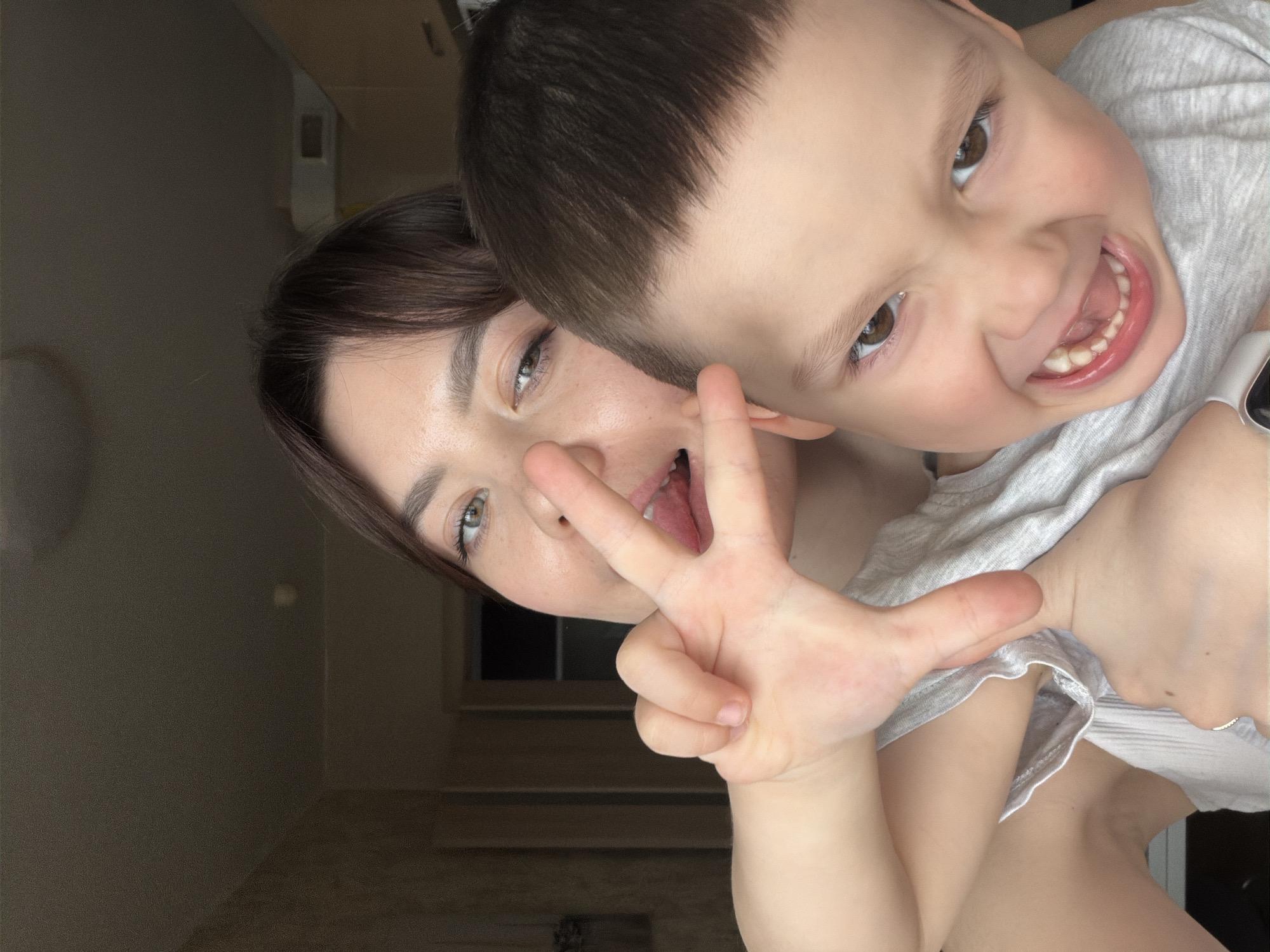A selfie shows a woman and a child indoors. The woman, with short dark hair, holds up a hand making a peace sign. Her mouth is open. The child, nestled behind her, is smiling broadly, showing their teeth. Both are wearing light gray t-shirts. The background includes a wall, a window, and hints of a staircase. The location is Chaikovsky, Russia.