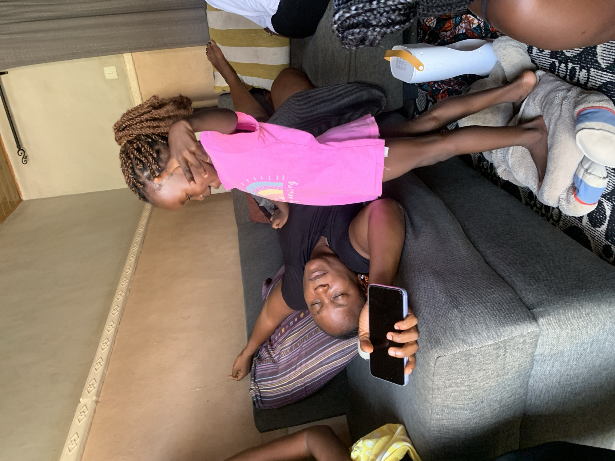 In Sabon Tasha, Nigeria, a woman is lying on her back on a grey couch, eyes closed, with a smartphone in her hand. A young girl, wearing a pink t-shirt with a rainbow graphic and the words "Be Amazing," stands next to the couch, facing the woman. The girl is playfully holding her hand up to her forehead, as if shielding her eyes or posing. The couch is adorned with a purple and white striped pillow. In the background, a textured wall with a decorative molding runs along the top. A white and yellow striped cushion is visible on the couch behind the woman. To the right, a white cylindrical object with a yellow handle, possibly a speaker or air freshener, rests on a patterned rug. The scene appears to be an indoor setting within a home.