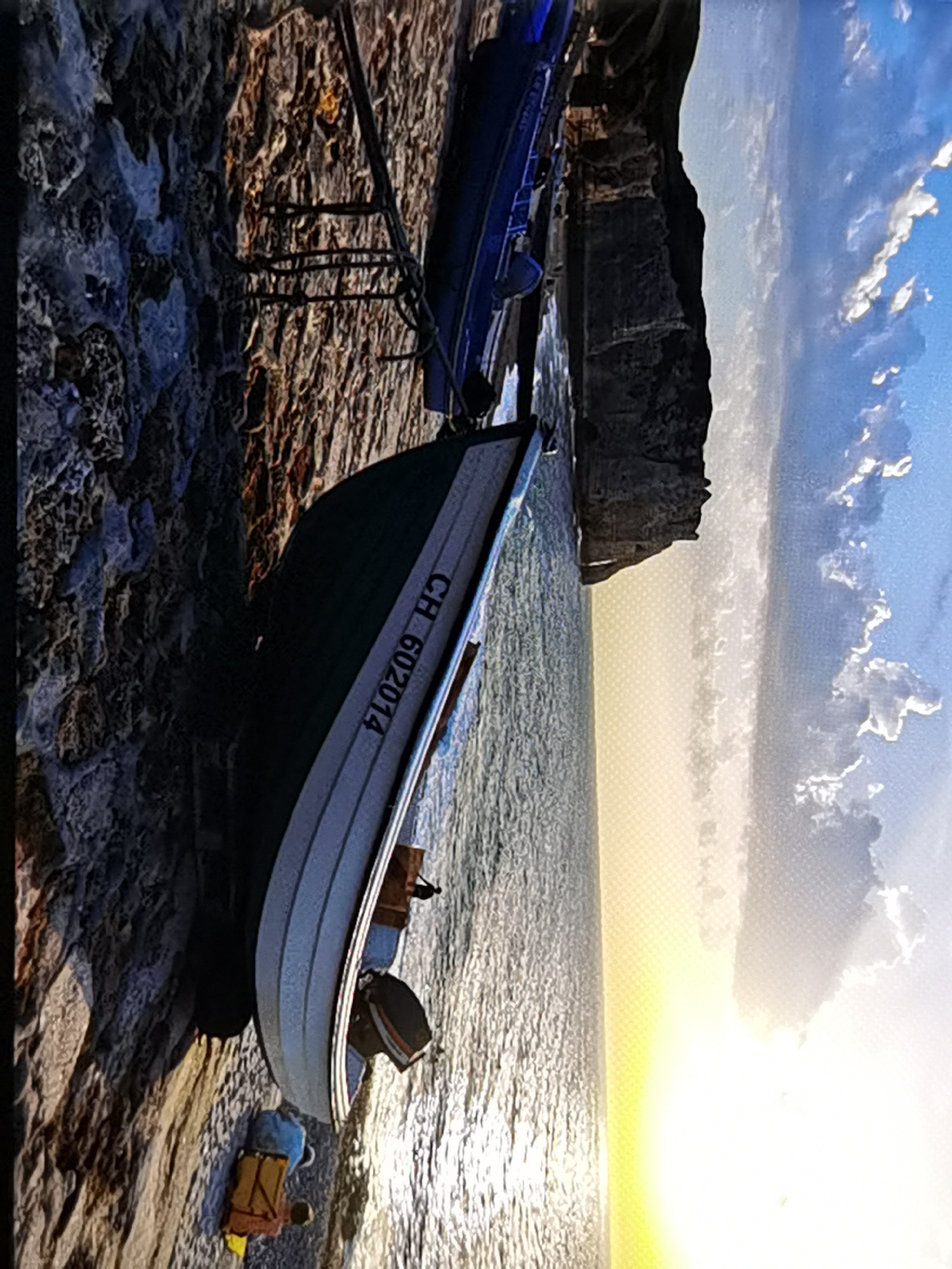 The image displays a tranquil scene at the base of the cliffs of Étretat, France. A small boat, identified by the markings "CH 602014," rests on the rocky shoreline. The boat is partially submerged in the calm, shimmering water, suggesting it has been pulled ashore or is waiting for the tide. Beside the boat, there are two bags, one blue and one yellow, suggesting a recent presence or belongings left behind. In the background, the iconic white cliffs of Étretat rise majestically, with a clear blue sky dotted with soft clouds overhead. The sunlight, low in the sky, casts a warm glow over the scene, creating a serene and picturesque atmosphere characteristic of this renowned coastal location.