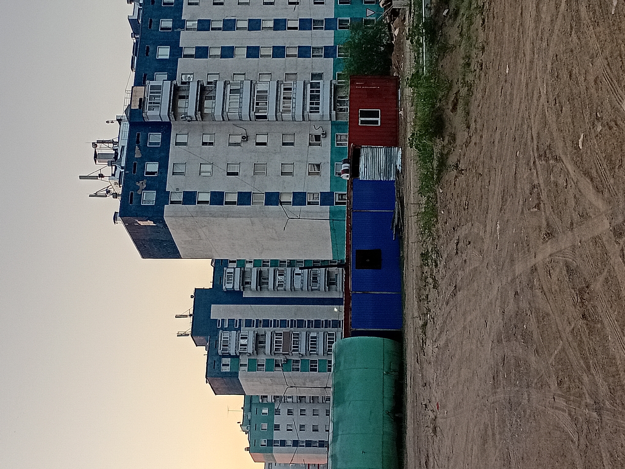 The image depicts a construction site in the city of Semey, Kazakhstan. In the background, several multi-story residential buildings with a blue and white facade are visible. In the foreground, there are temporary structures including a large green cylindrical tank, a blue shipping container, and a red container with a single window. The ground is unpaved and shows signs of tire tracks, indicative of ongoing construction activity. The sky is a pale orange, suggesting either sunrise or sunset. No people are visible in the frame.