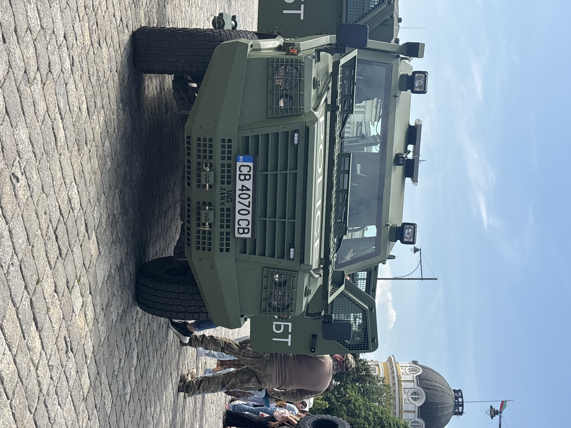 The image shows a green military vehicle parked on a cobblestone street. The vehicle has a Bulgarian license plate that reads "CB 4070 CB". "POLICE" is displayed on the windshield. People are standing around the vehicle, one of whom appears to be wearing camouflage clothing. In the background, there is a dome-shaped building and a Bulgarian flag. The location is in Sofia, Bulgaria, likely near the St. Alexander Nevsky Cathedral, given the visible architectural style.