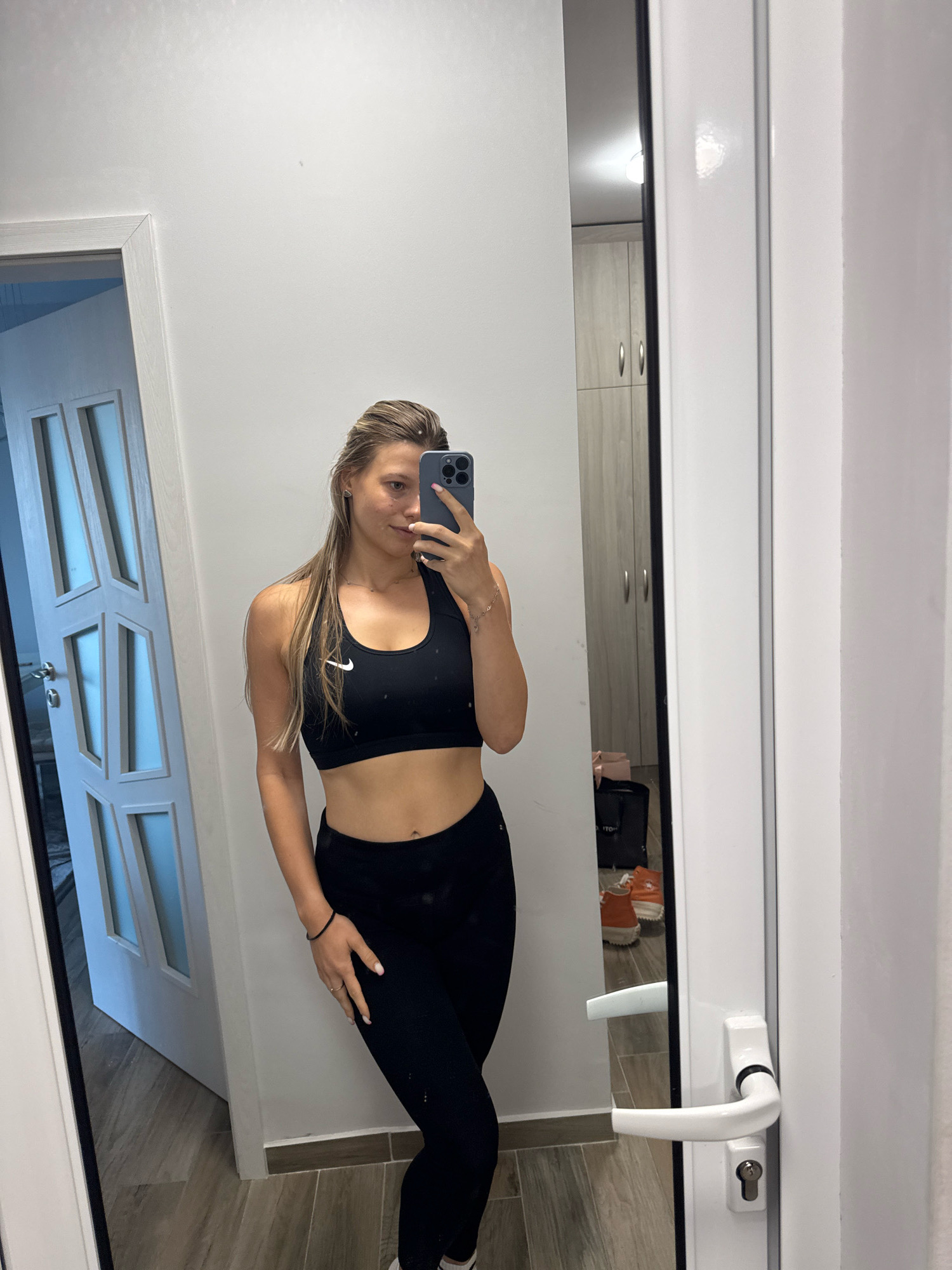 A woman is taking a mirror selfie in what appears to be a domestic setting, possibly a hallway or a dressing room. She is wearing a black Nike sports bra and black leggings. Her hair is long and blonde, pulled back in a ponytail. She is holding a smartphone in her right hand, angled to capture the reflection.
The background is mostly white walls. To the left, there is a white door with frosted glass panels. To the right, a large mirror reflects the woman and a portion of the room behind her. In the reflection, we can see a closet with light wood doors, and on the floor, there are a black bag and a pair of orange sneakers. The floor appears to be tiled with a wood-grain pattern.
The image does not contain any visual information that would allow for a specific location in Sofia, Bulgaria.
