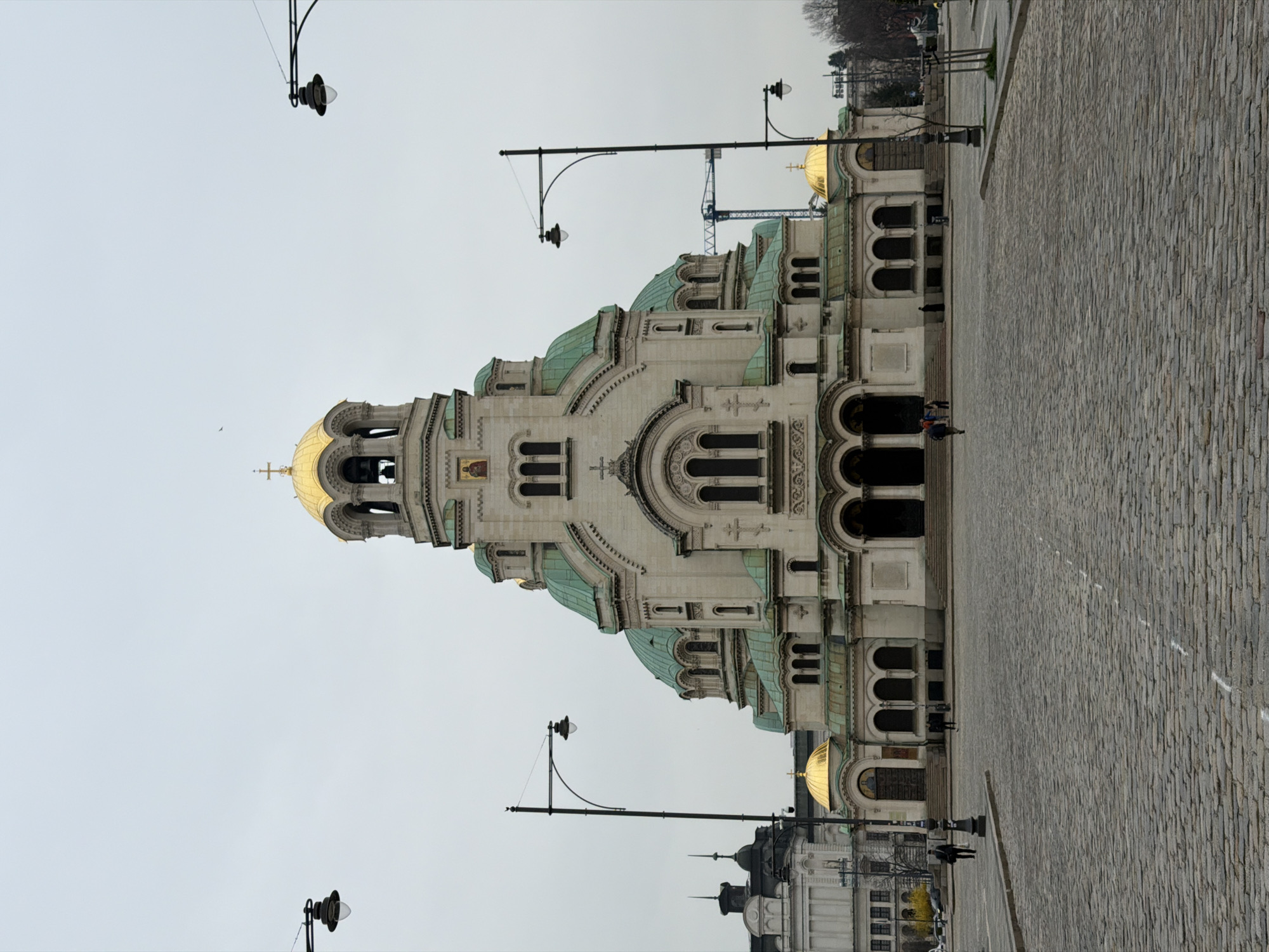 The image depicts the Alexander Nevsky Cathedral in Sofia, Bulgaria, from a slightly low angle. The cathedral's facade is white stone with green copper domes and gilded gold domes. The foreground is a paved cobblestone square. A few people are present near the base of the cathedral. Streetlights with curved arms are visible. The sky is overcast. A blue construction crane is seen in the background.