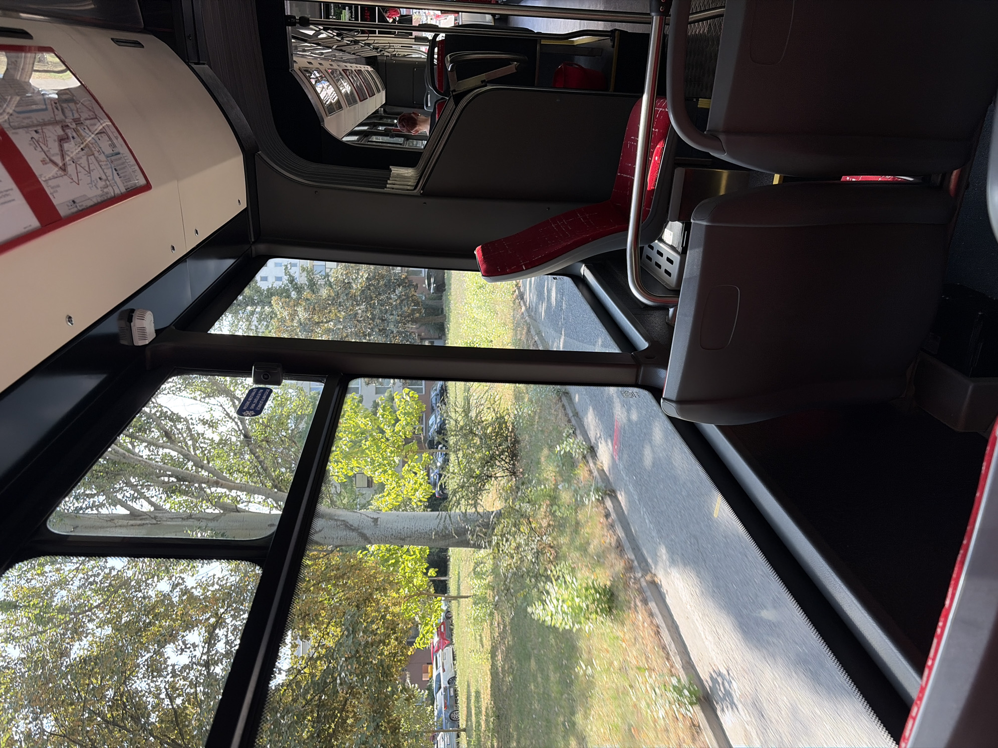 The image depicts the interior of a public transport vehicle, likely a bus, with a view out of the window. The viewpoint is from a seat on the right side of the bus, looking towards the left and slightly forward. Outside the window, a sunny scene shows trees, green grass, and parked cars, suggesting a park or residential area. The foreground inside the bus features dark-colored seats and metal handrails. In the background, more seats are visible, along with a person with short, light-colored hair seated further down the aisle. A map is displayed on the ceiling of the bus. The overall impression is that of a typical urban public transport environment on a clear day. The presence of a map with a route and destination indicators, along with the general design of the bus interior, strongly suggests this is within the public transport system of Nitra, Slovakia, given the context provided.