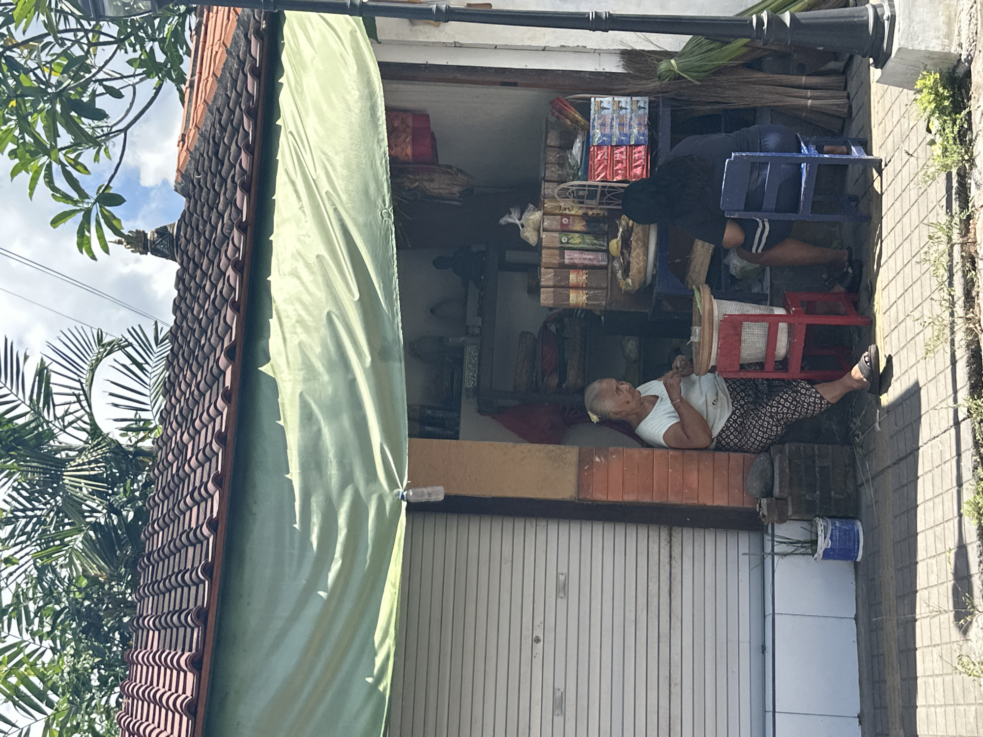 Two women are seated outside a small shop in Ubud District, Indonesia, under a green tarp that extends over the storefront. The woman on the left, appearing older, is wearing a light blue top and a patterned skirt. She is seated on a red chair and appears to be engaged in an activity, possibly eating or preparing food, with a small bowl on a table in front of her. The woman on the right, younger and with dark hair, wears a dark t-shirt and striped shorts. She is seated on a blue chair and faces away from the camera. The shop itself displays various items on shelves, including packaged goods, what appear to be incense sticks, and decorative fabrics. A rolling metal door is visible to the left of the shop entrance. The ground is paved with interlocking square tiles. Overhead, palm fronds and other tropical foliage are prominent, with a glimpse of a cloudy sky. A decorative element, possibly a shrine or finial, is visible on the roof of the building.