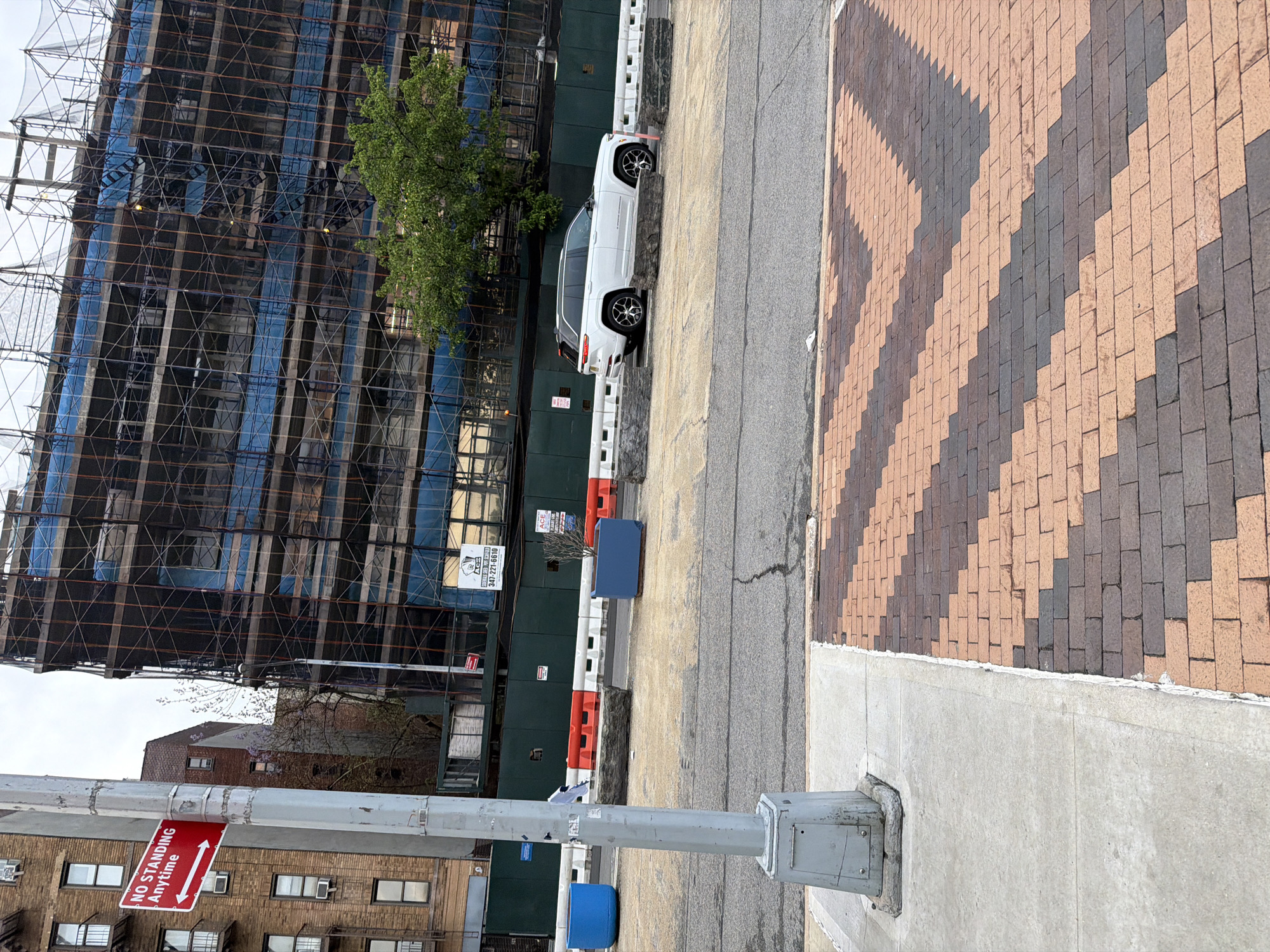 The scene shows a street in New York City, United States, with a construction site dominating the view. A tall building is under construction, encased in scaffolding and blue safety netting. A white SUV is parked along the curb, next to a section of sidewalk and a brick-paved area featuring a herringbone pattern. A street sign reading "No Standing Anytime" is visible, attached to a gray metal pole. Other buildings are seen in the background, as well as blue construction barricades. The overall impression is an urban environment with ongoing construction.