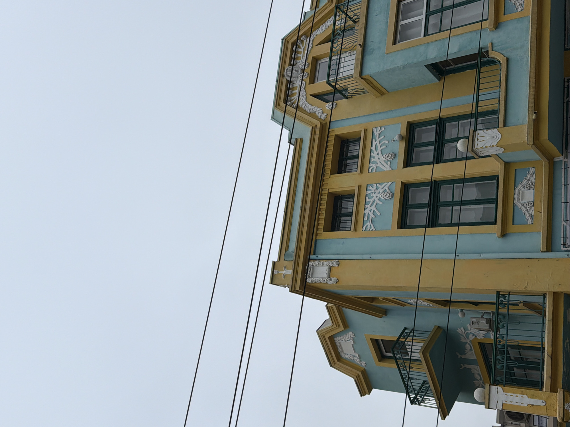 A low-angle shot of a building's corner facade in Varna, Bulgaria. The building features a decorative architectural style with light blue and yellow paint. Windows are framed in dark green, with white decorative plaster elements between them. Small wrought-iron balconies are present. Electrical wires run across the left side of the frame against a light gray sky.