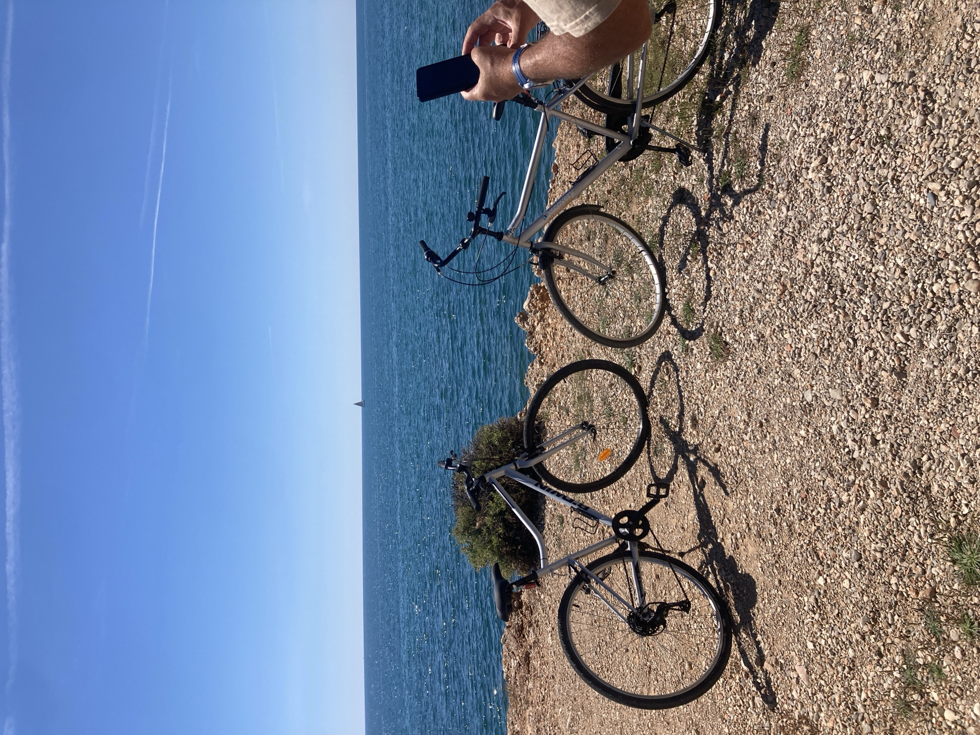 The image shows two bicycles parked on a rocky cliff overlooking the sea. One person is holding a smartphone, presumably taking a photo. The sky is clear and blue. A sailboat is visible in the distance. The location is unknown.