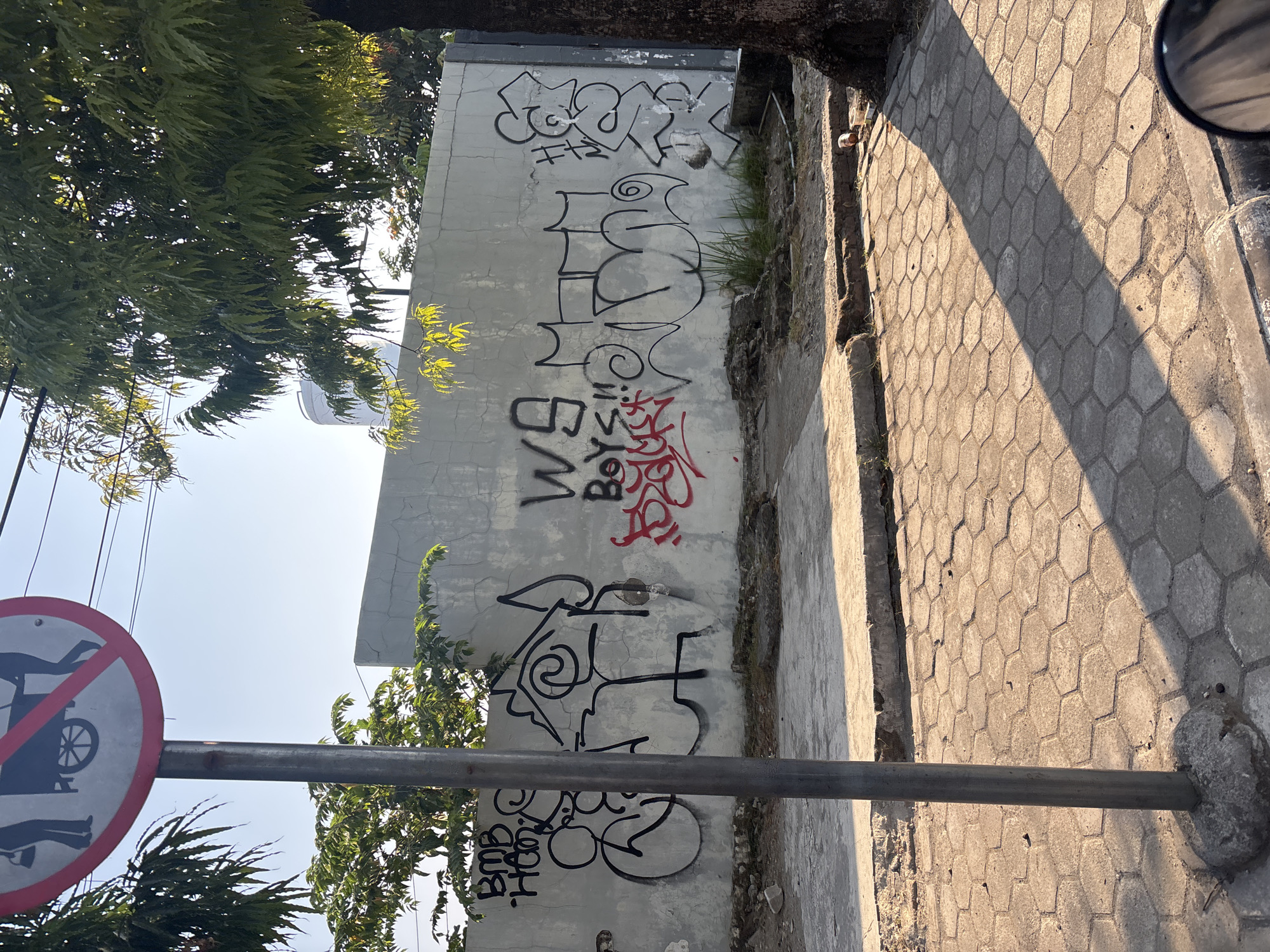 The image depicts a street scene in Cirebon, Indonesia, dominated by a concrete wall covered in black and red graffiti. A gray pole stands in the foreground, supporting a circular red and white sign prohibiting the pushing of carts. To the left of the pole, the graffiti includes what appears to be the letters "BMB HAM". In the center, a stylized house-like structure is drawn, with swirls and angular lines. Further to the right, black graffiti reads "WS BOYS!!!" and "MCK". Red graffiti is also visible on the wall, though its content is obscured. The wall itself shows signs of age and cracking. Behind the wall, a white water tank and trees are visible against a clear blue sky. The ground in front of the wall is paved with hexagonal gray stones, with some debris and overgrown grass. Sunlight casts long shadows across the pavement, indicating it is either early morning or late afternoon. A large tree trunk is visible on the far right of the image.