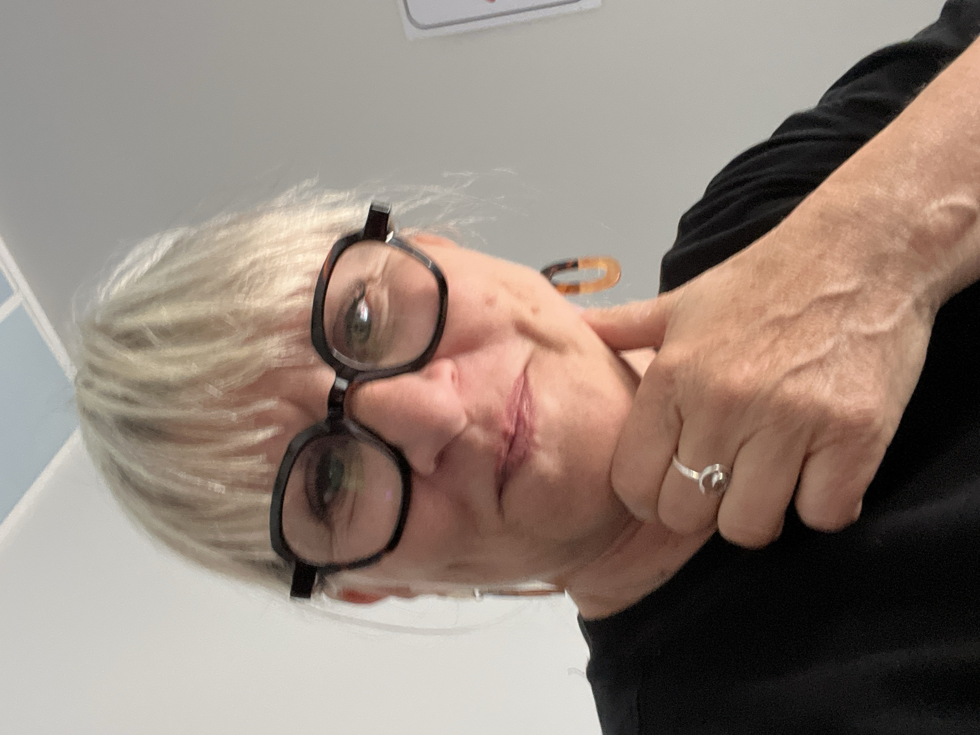 An extreme close-up portrait, rotated 90 degrees clockwise, captures a person, likely a woman, from a low-angle perspective. The subject has short, wispy blonde or white hair and wears thick, black-framed glasses that obscure part of her eyes. Her head is tilted upwards, and a prominent vertical scar is visible on her upper lip/philtrum area. Her right hand is raised, with fingers resting gently against her neck or chin. Veins are visible on the back of her hand, and she wears a silver ring, possibly shaped like a horseshoe or two interlocking C's, on her ring finger. She is dressed in a black top or shirt, visible on the right side of the frame. The background is a plain, light grey or white wall, with a small section of light blue visible in the upper left corner, suggesting an indoor setting. The person's expression appears neutral or slightly thoughtful as she looks towards the top of the frame. No text is visible. The image provides no specific time of day or weather cues beyond an indoor environment, which is situated in Saint-Galmier, France, as per the context.