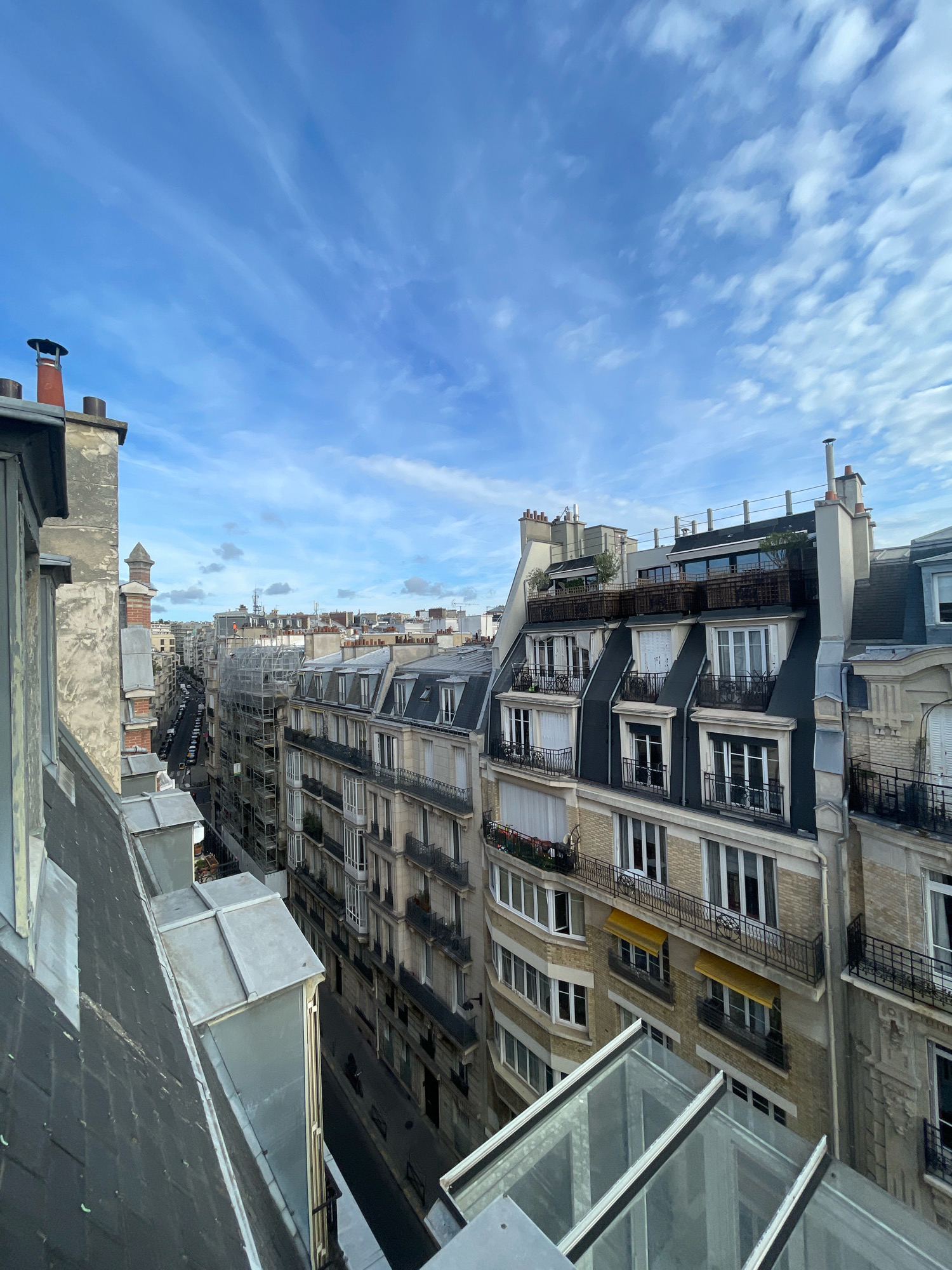 The image is a high-angle, eye-level shot of a Parisian street scene, likely taken from a rooftop or a higher floor of a building. The view looks down a narrow street lined with Haussmann-style apartment buildings. These buildings feature cream-colored stone facades, mansard roofs with dormer windows, and ornate iron balconies. Many windows are visible, some with yellow awnings extended. A street with parked cars is visible below, and scaffolding is present on one of the buildings. The sky above is a bright blue with scattered white clouds, suggesting fair weather. In the foreground, the edge of a dark, sloped roof with metal ductwork is visible, as are sections of glass skylights. The overall impression is an urban panorama of Paris, characterized by its distinctive architectural style.