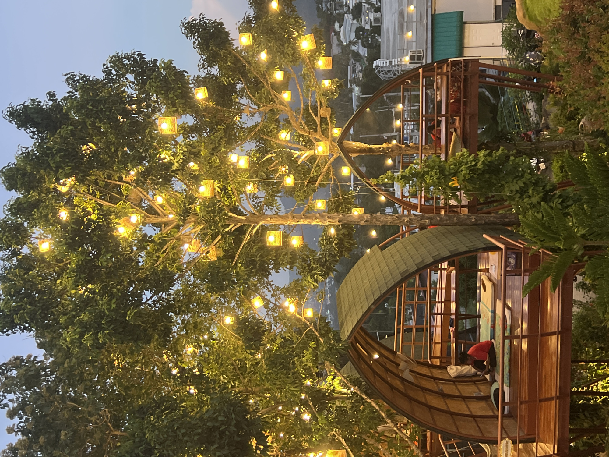 The image depicts an outdoor seating area at dusk in Ko Samui District, Thailand. Two elevated wooden platforms, one with a curved, thatched roof resembling a hobbit house, are nestled among trees adorned with numerous glowing yellow lanterns. The platforms offer views of the surrounding cityscape, with buildings and lights visible in the distance. One platform is occupied by a person sitting on a mat, wearing a red shirt, and looking at a phone. Another person is seated at a table on the second platform. Lush greenery and foliage surround the seating areas. The sky is a soft blue, indicating the transition from day to night.