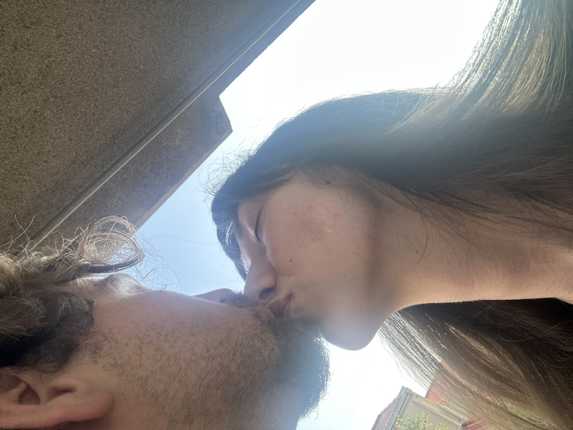 A close-up, low-angle shot captures two individuals embracing in a kiss. The person on the left, partially visible, has a beard and is angled towards the person on the right. The person on the right, a female, has her eyes closed and her lips pursed. Her long, brown hair frames her face, and the sunlight creates a lens flare effect. In the background, a bright sky and the corner of a building can be seen. The image is shot from below, looking up at the couple. The context of Belgrade, Serbia, is not discernible from the visual information provided in the image.