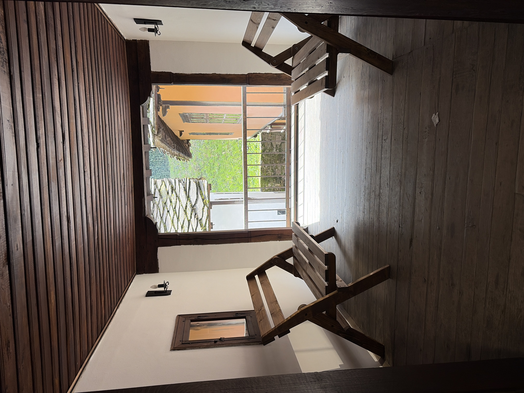 A view from an interior space in Gabrovo, Bulgaria reveals a room with a combination of white and brown wood-paneled walls and a wooden floor. Light fixtures are mounted on the walls. Two wooden folding chairs are positioned in the room. Outside, a garden view with trees and a stone wall is visible through a doorway.