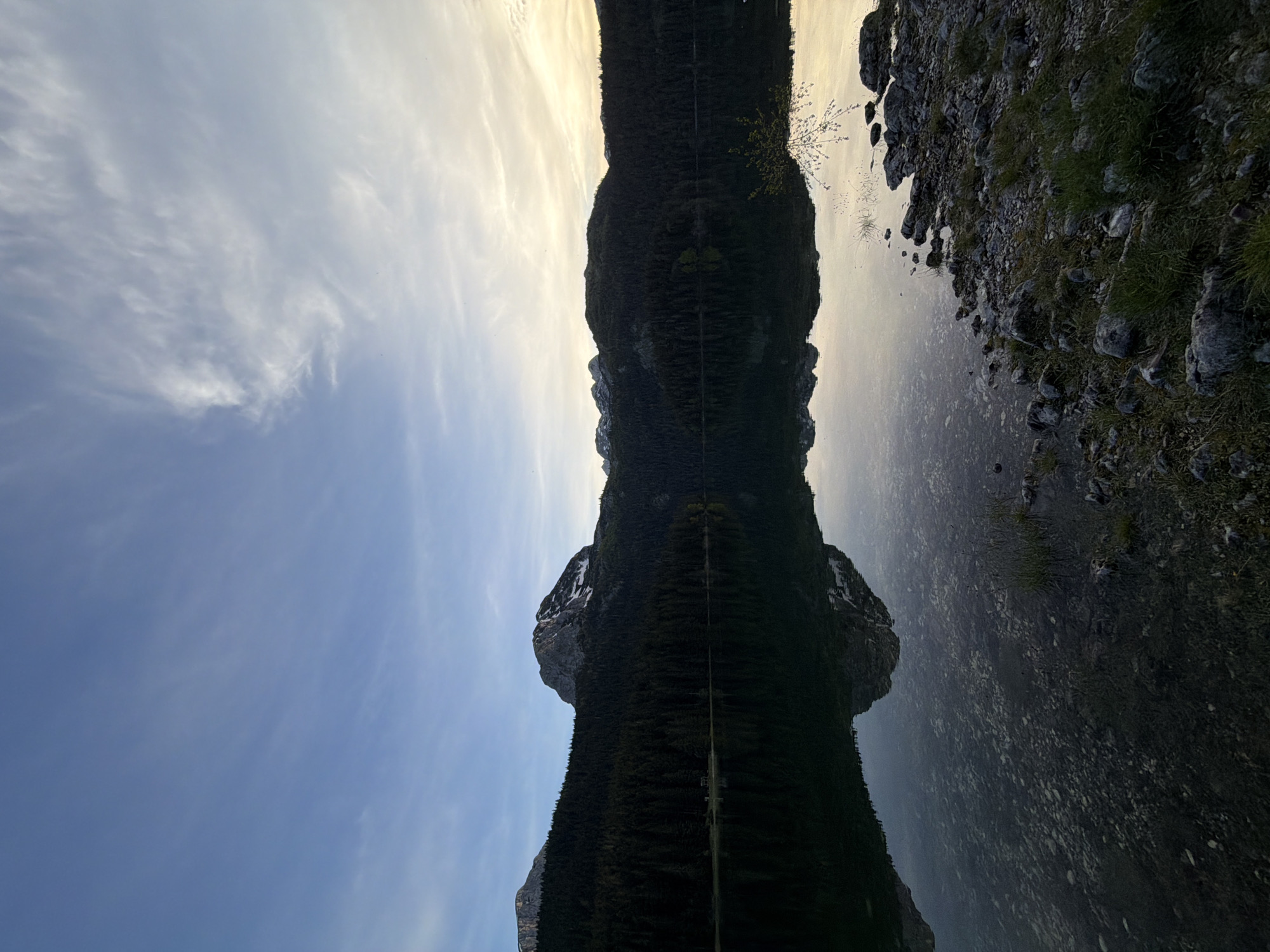 The image depicts a landscape scene, mirrored vertically. The upper half shows a sky with wispy clouds and a blue hue transitioning to lighter tones near the center. The lower half appears to be a reflection in water.
In the center, a dark, elongated mass stretches vertically, resembling a mountain range covered in trees. There's a clear line dividing the mountain and its reflection. On the right side, the land meets the water with rocks and grass. The mirrored reflection of these rocks and grass gives texture to the reflection. This scene appears to be a view of the Durmitor National Park, located in Pitomine, Montenegro, known for its mountains and lakes. There are no people visible in the image.