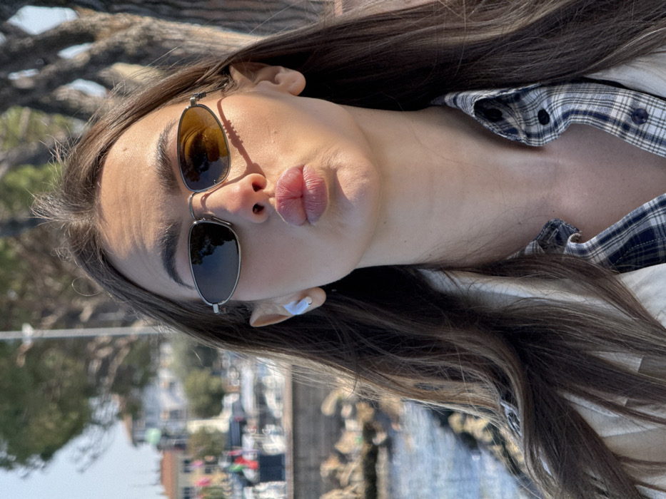 A close-up photograph depicts a female subject outdoors in Peschiera del Garda, Italy. The subject has long brown hair and wears dark, oval-frame sunglasses, with the right lens reflecting an amber light. Her lips are pursed forward. A white wireless earbud is present in her left ear. She is dressed in a light-colored outer garment over a blue and white plaid shirt. The blurred background features a body of water with moored boats, distant buildings with light facades and red roofs, and sparse trees. The scene is brightly lit by direct sunlight.