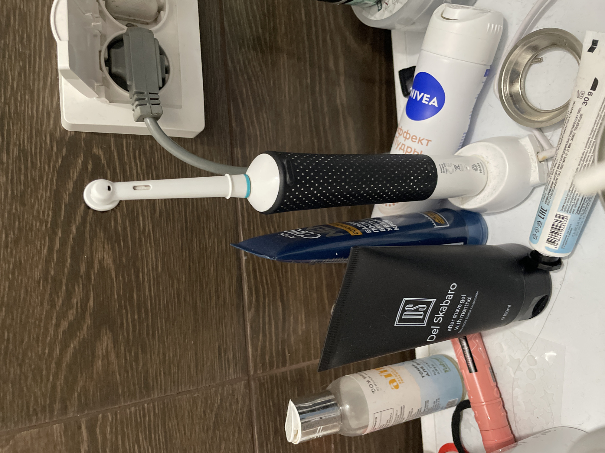 The image depicts a close-up, overhead view of a bathroom countertop in Sinyavino, Russia. The countertop is cluttered with various personal hygiene products. A white electric toothbrush with a black patterned grip is plugged into a white wall socket, its gray cord extending to the charger base. To the left of the toothbrush, a clear plastic bottle with a silver cap and a label that appears to read "DOM TRI" and "айн" is visible. Next to it is a black tube labeled "Del Skabaro after shave gel with menthol." A blue tube, possibly a toiletry product, is positioned behind the black tube. In the foreground, a pink object with a silver metallic tip lies horizontally, and to its right, a white tube with a blue and white logo that reads "NIVEA" is present. Further to the right, a metal ring, possibly part of a sink fixture, rests on the countertop. The background consists of a brown tiled wall.