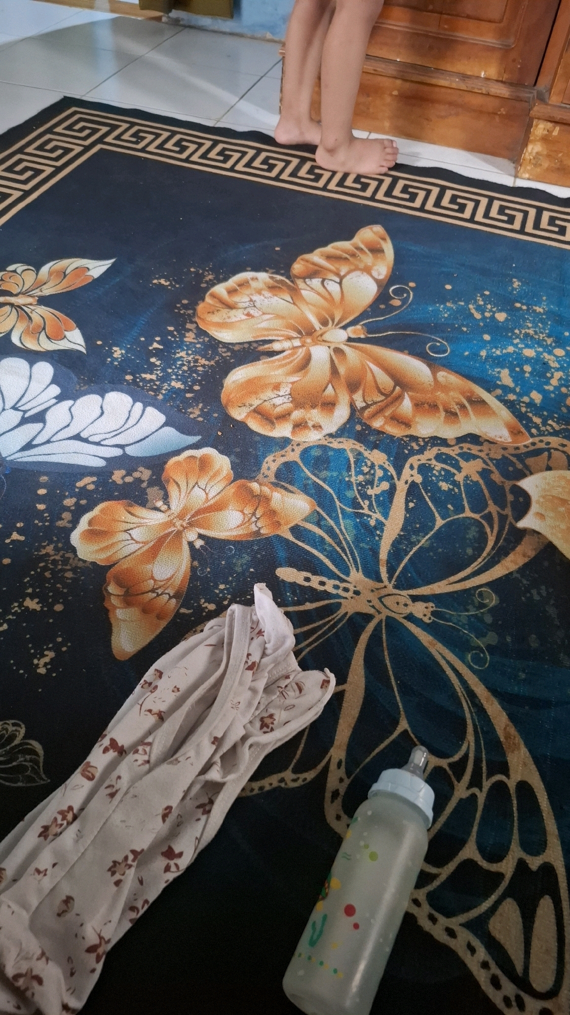 A child's bare feet stand on the edge of a dark blue rug with a golden butterfly and border pattern. To the left, a light-colored garment with a floral pattern is folded. To the right, a baby bottle with a blue cap and a partially visible cartoon image is positioned on the rug. The background shows a wooden surface and white tiled flooring. The scene appears to be in a domestic setting, possibly a home in Bandar Lampung, Indonesia.