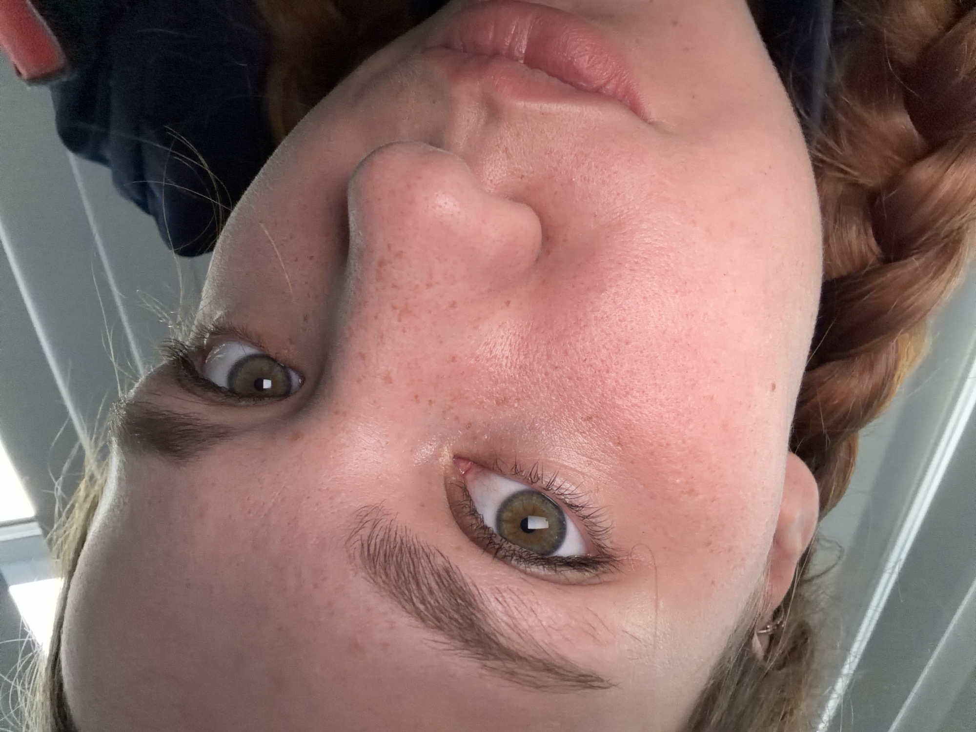A close-up, low-angle, medium shot shows a young woman's face from the nose up, angled slightly to her left. Her skin is fair with scattered freckles across her cheeks and nose, and a faint blush. Her left eye, which is in focus, is a hazel-green with long eyelashes and a defined brown eyebrow. Her right eye is partially visible, and also appears hazel-green. Her lips are slightly parted, revealing a hint of her teeth. She has a braid of auburn hair resting on her left shoulder. The background is blurred and appears to be an indoor setting with vertical white blinds or partitions. The lighting is soft and natural.