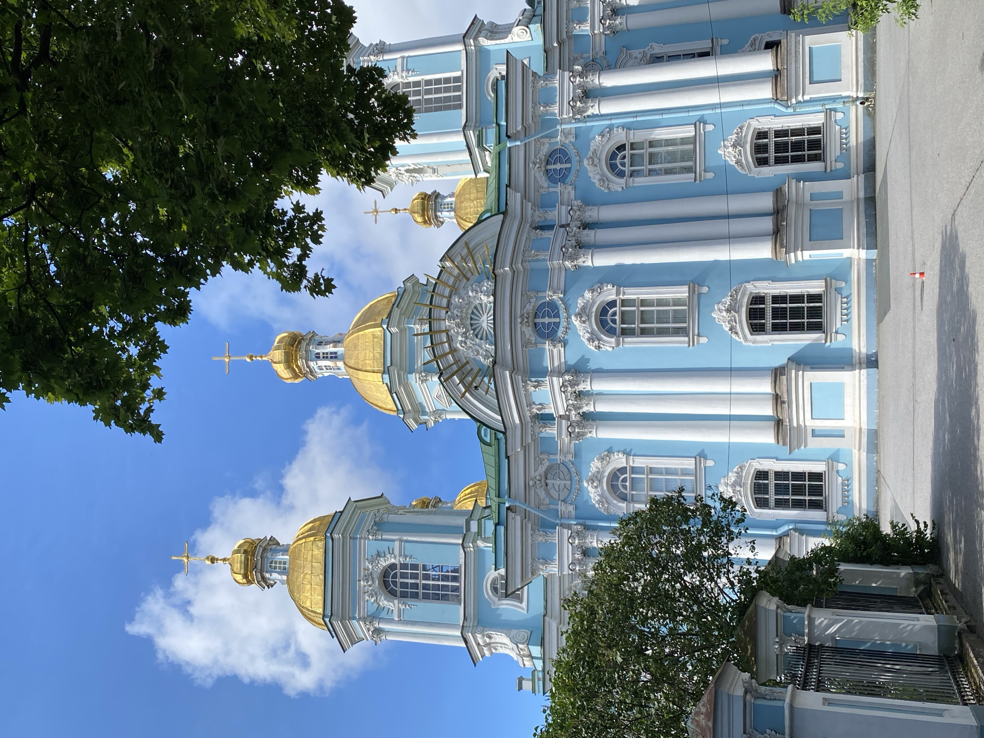 The image shows the blue facade of St. Nicholas Naval Cathedral in Saint Petersburg, Russia, on a clear day with a blue sky and scattered clouds. The cathedral features multiple golden domes with crosses, ornate white detailing, and large windows. Green trees partially obscure the view of the building on the right side of the frame, and some foliage is visible on the left. A black metal fence with decorative pillars lines the foreground on the left. The ground in front of the cathedral is paved.