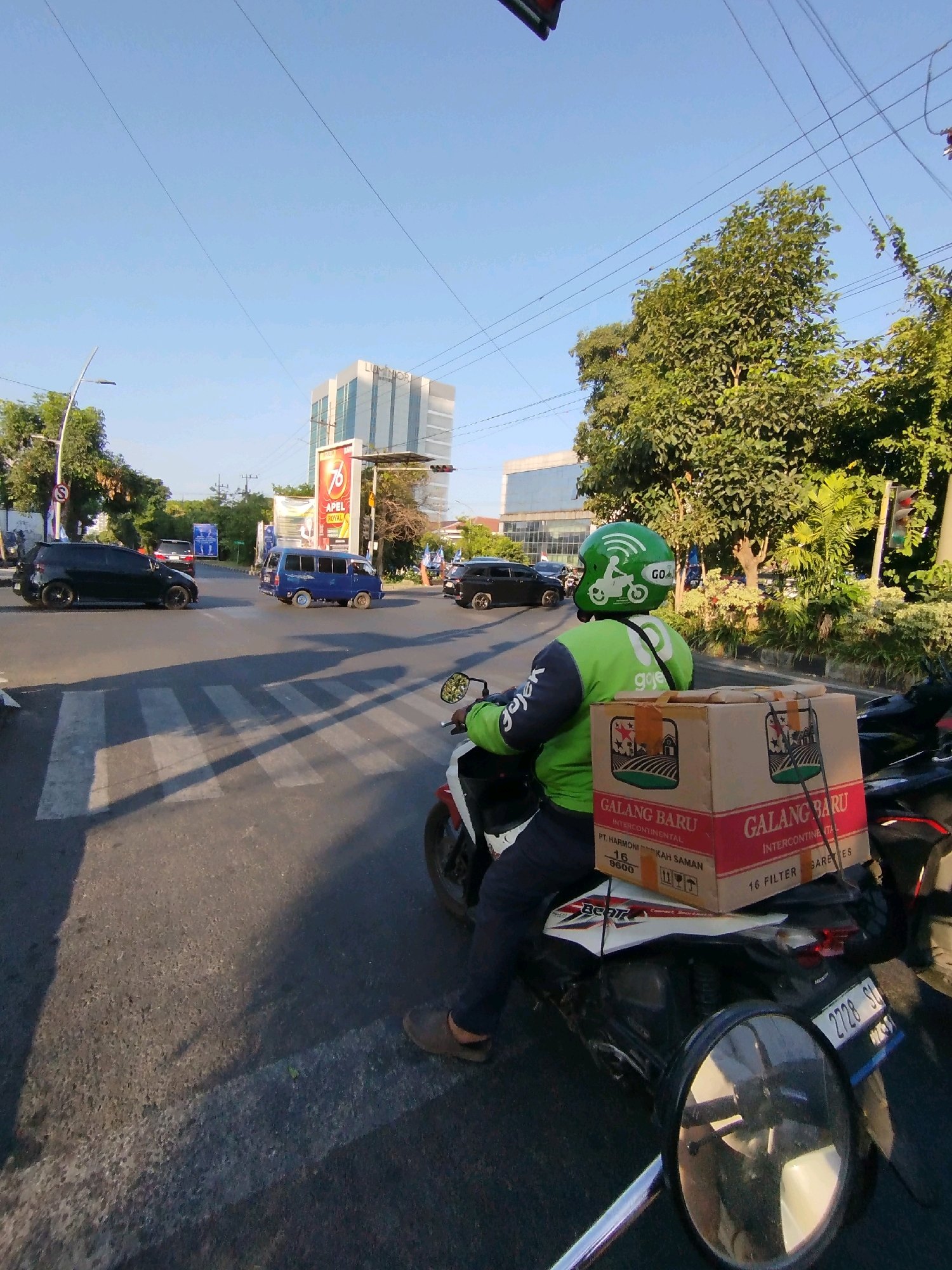 A male delivery driver wearing a green helmet with a "GoGo" logo and a green jacket is riding a scooter across a pedestrian crossing. The scooter is carrying a large cardboard box on the back, labeled "GALANG BARU INTERCONTINENTAL". The background shows a street intersection with several cars and a blue van. In the distance, a modern building with glass windows is visible, along with trees and a clear blue sky. The scene appears to be in Surabaya, Indonesia, indicated by the "GoGo" logo on the driver's gear, a popular ride-hailing service in the country, and the general urban streetscape. The license plate of the scooter is partially visible, showing "2728 SU".