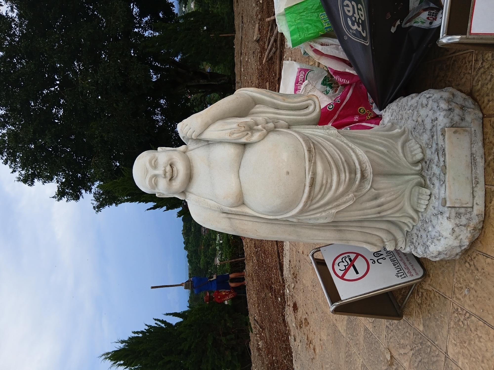 A large, white marble statue of a smiling, corpulent Buddha is positioned in the foreground, facing the viewer. The statue is on a rough, light-colored stone base, which in turn rests on a tiled surface. To the left of the Buddha statue, a white and silver sign with a red circle and diagonal line over a smoking cigarette indicates a no-smoking zone, with Thai text below. Behind the statue, the ground is covered with reddish-brown soil and scattered debris, suggesting a construction or work area.
In the middle ground, a person wearing a blue shirt and dark shorts is standing and holding a long wooden pole upright. To the left of this person, a smaller figure, possibly a scarecrow or a child, is wearing a red top and dark bottoms. The background reveals a lush green landscape with many trees, rolling hills, and some distant buildings. The sky is overcast with a pale blue hue, and scattered birds are visible. To the right of the Buddha statue, several large bags, some brightly colored, are piled up, and a black umbrella lies on the ground. The overall setting appears to be an outdoor location in Baan Lao Phattana, Thailand, possibly a temple or a public area under development.